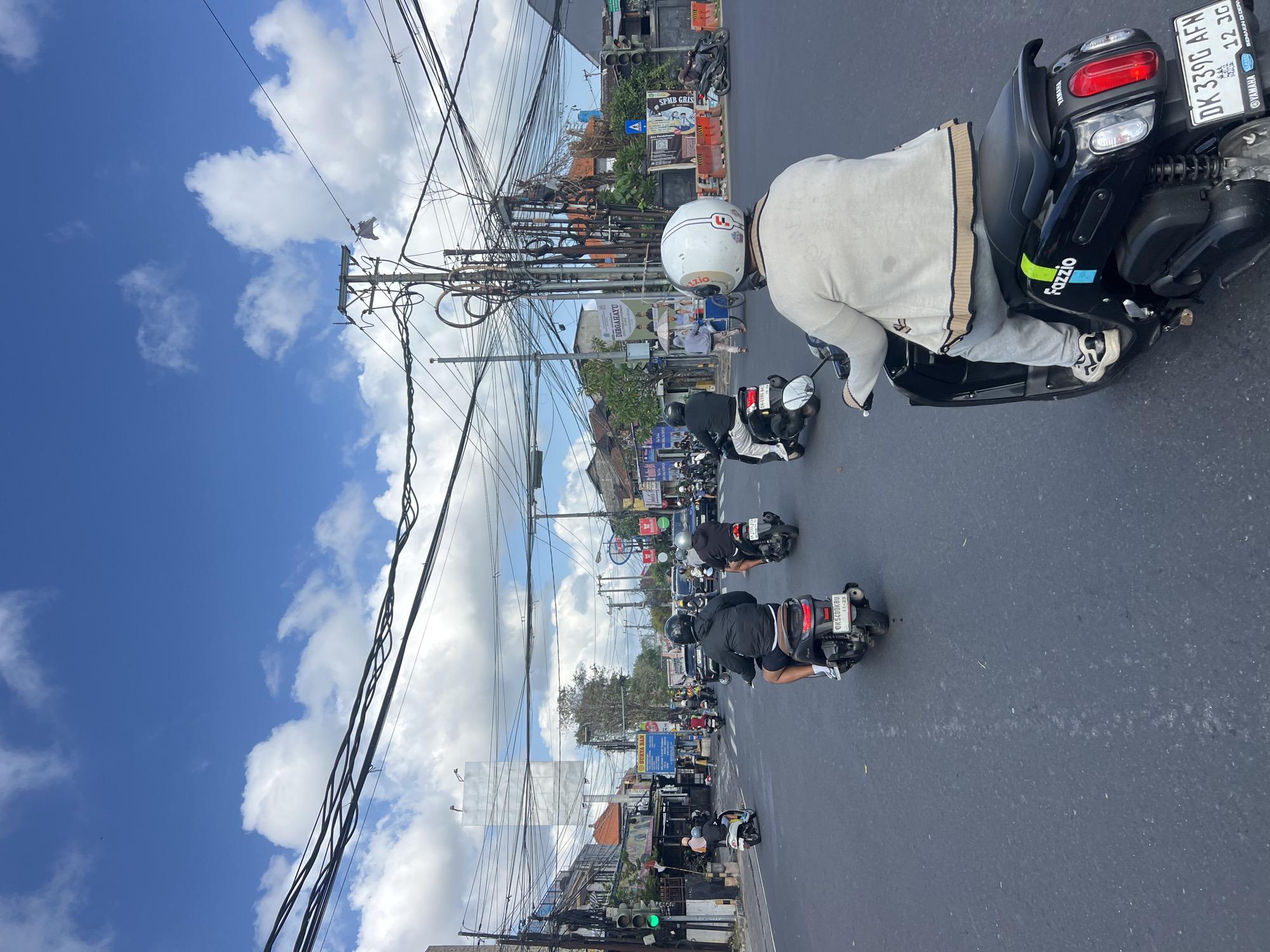 This is a street-level, medium shot captured in Denpasar, Indonesia, on a day with a partly cloudy blue sky. The image is dominated by a dense network of overhead power lines and utility cables, a common sight in the city. In the foreground, the back of a black Fazzio Yamaha scooter is visible, ridden by a person wearing a white helmet and a light-colored sweater over gray sweatpants. The scooter's license plate reads "DK 339G AFN".
The street is busy with traffic, primarily scooters and motorcycles. Several other riders are visible, some in the immediate vicinity and others further down the road. One rider in front of the Fazzio scooter wears a black helmet and shorts. To the left, a white scooter is also present. In the mid-ground, a traffic light shows a green signal. Buildings line the street, some appearing to be shops or businesses with signs. Orange traffic barriers are also present on the right side of the road. In the distance, taller buildings are visible, including what appears to be a blank billboard structure. The overall scene depicts a typical bustling intersection in Denpasar, characterized by heavy traffic and extensive electrical infrastructure.