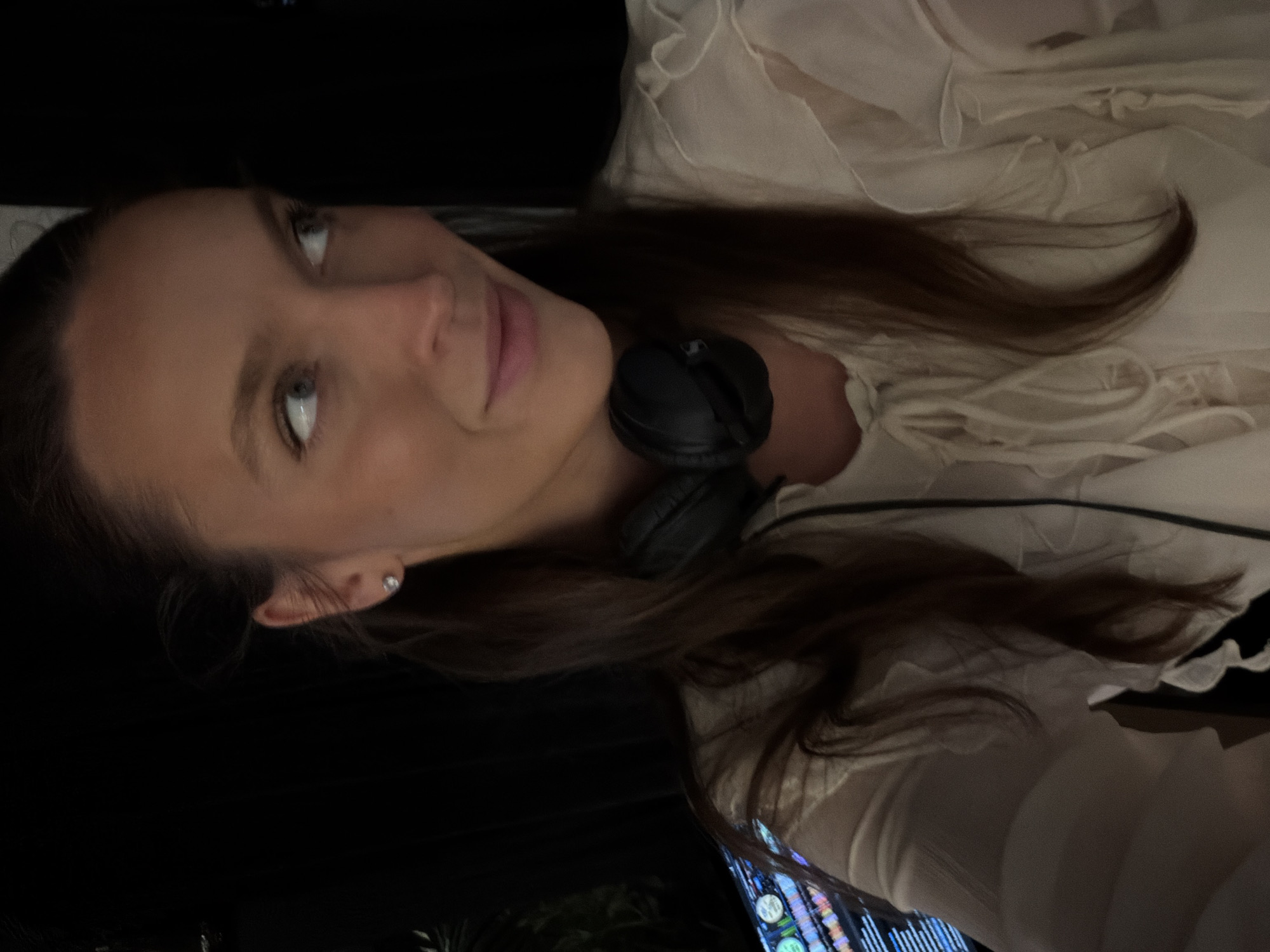 A woman with long brown hair and blue eyes is wearing a white ruffled shirt and black headphones. She is looking at the camera and smiling slightly. She has a small earring in her left ear. A screen is visible in the background. The location is not discernible from the image.