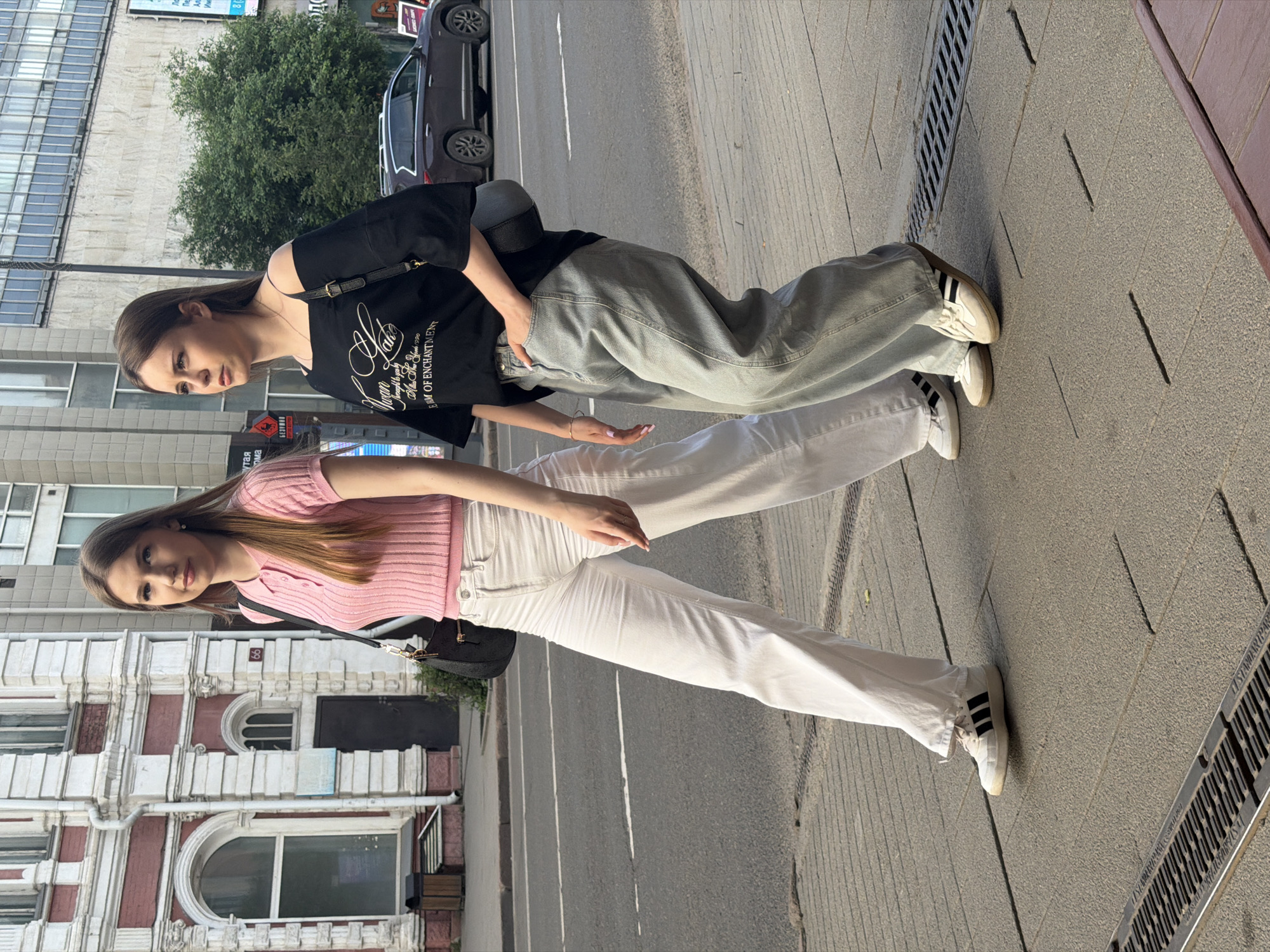 Two young women are standing on a sidewalk in a city. One woman on the left is wearing a black t-shirt with white text and light gray baggy jeans. She has a black handbag slung over her shoulder. The woman on the right is wearing a pink ribbed short-sleeve shirt and white pants. She also has a black handbag. Both women are wearing white sneakers with black stripes. The background shows a building with red and white accents, a street with a car, and some trees. The sunlight suggests it is daytime. The text on the black t-shirt is partially visible and appears to be in English, possibly mentioning "EUM OF ENCHANTMENT". The number "8" is visible on the building. The image appears to be taken in Krasnoyarsk, Russia. The women are posing for the photo, with one looking towards the camera and the other looking away.