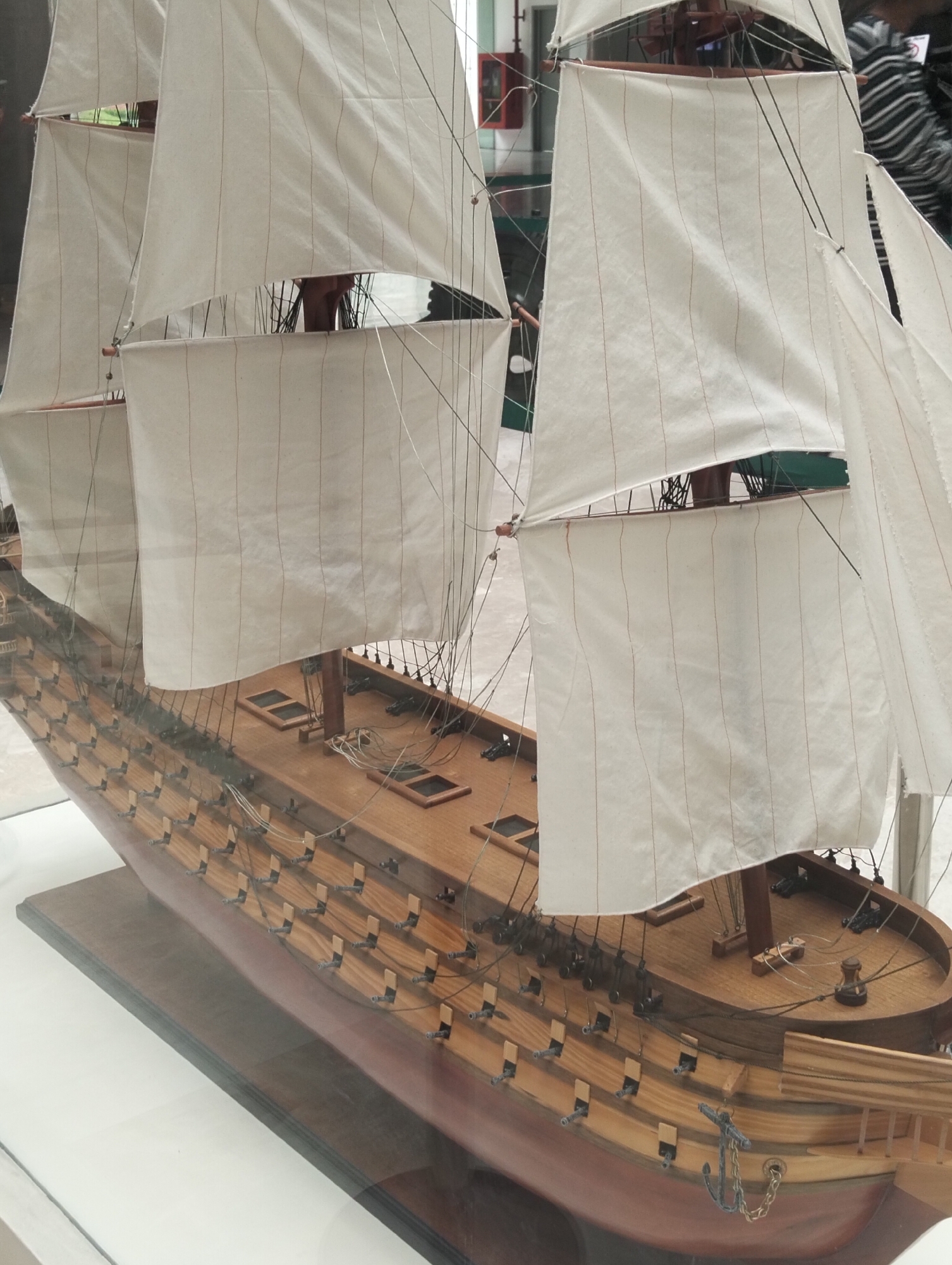 A model ship is encased in glass. The model ship is red and tan and has white sails. The model has many small, black cannons. The model ship also has an anchor and chain on its starboard bow. The model ship is likely in a museum in the city of Dhaka, Bangladesh.