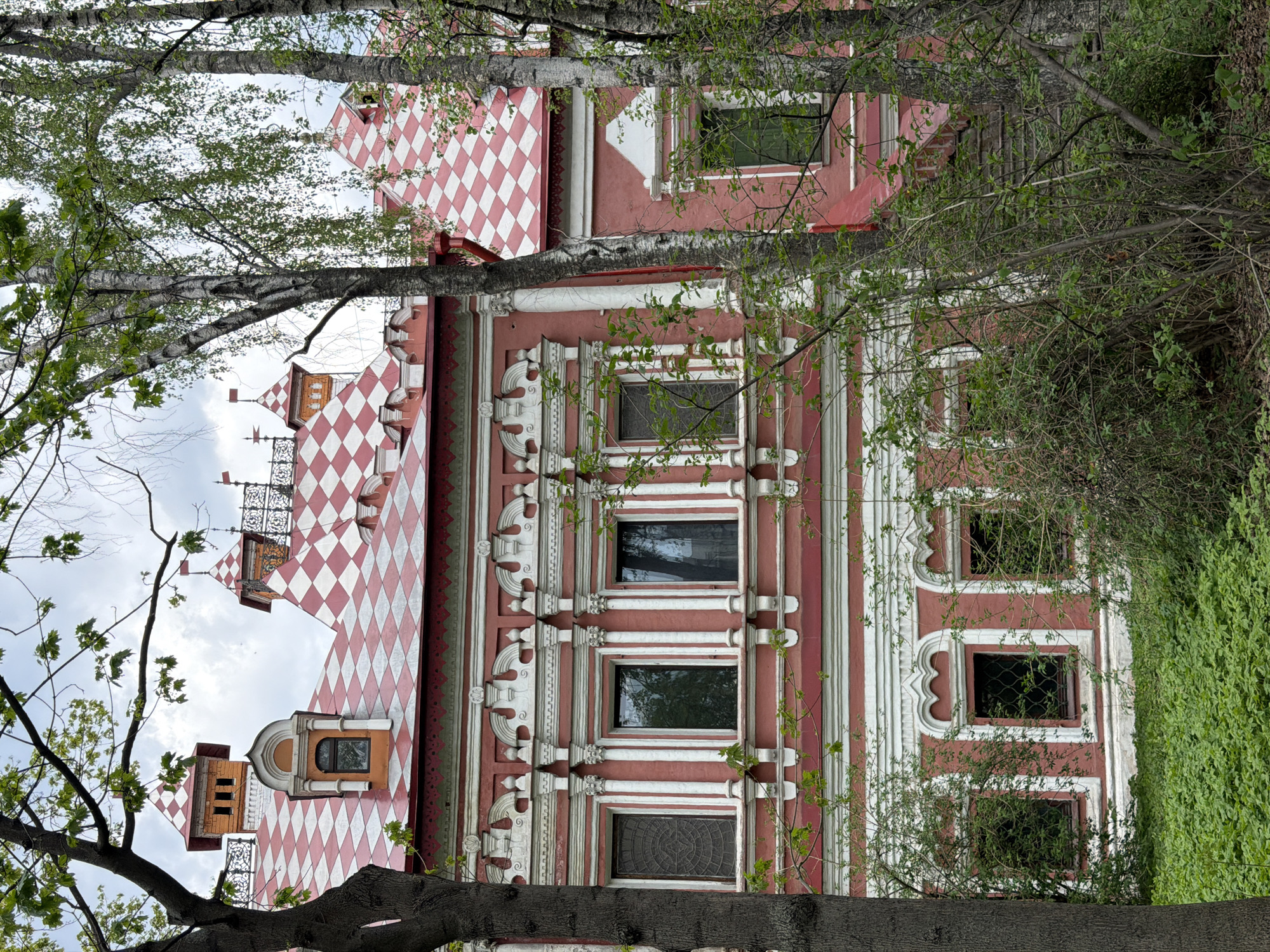 The image shows a portion of Saint Basil's Cathedral in Moscow, Russia, partially obscured by tree branches. The cathedral's distinctive red and white checkered roof and ornate facade are visible. The sky is cloudy. The trees are green.