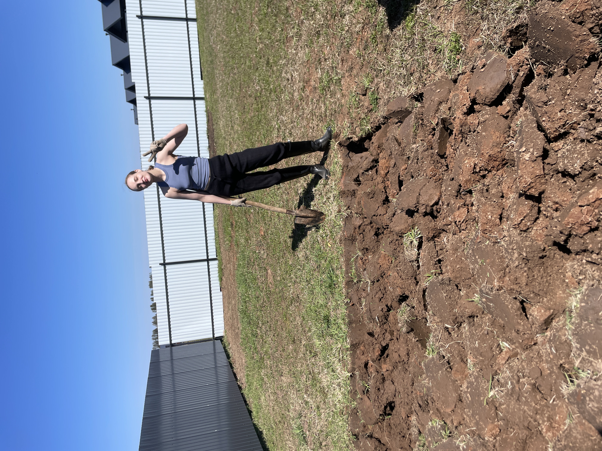 A young woman wearing a blue tank top, black pants, and boots is standing in a yard, holding a garden fork. She is making a peace sign with her gloved hand. The ground has been partially dug, revealing dark brown soil. A fence borders the yard, and the sky is blue. The location is Подшивалово, Russia.