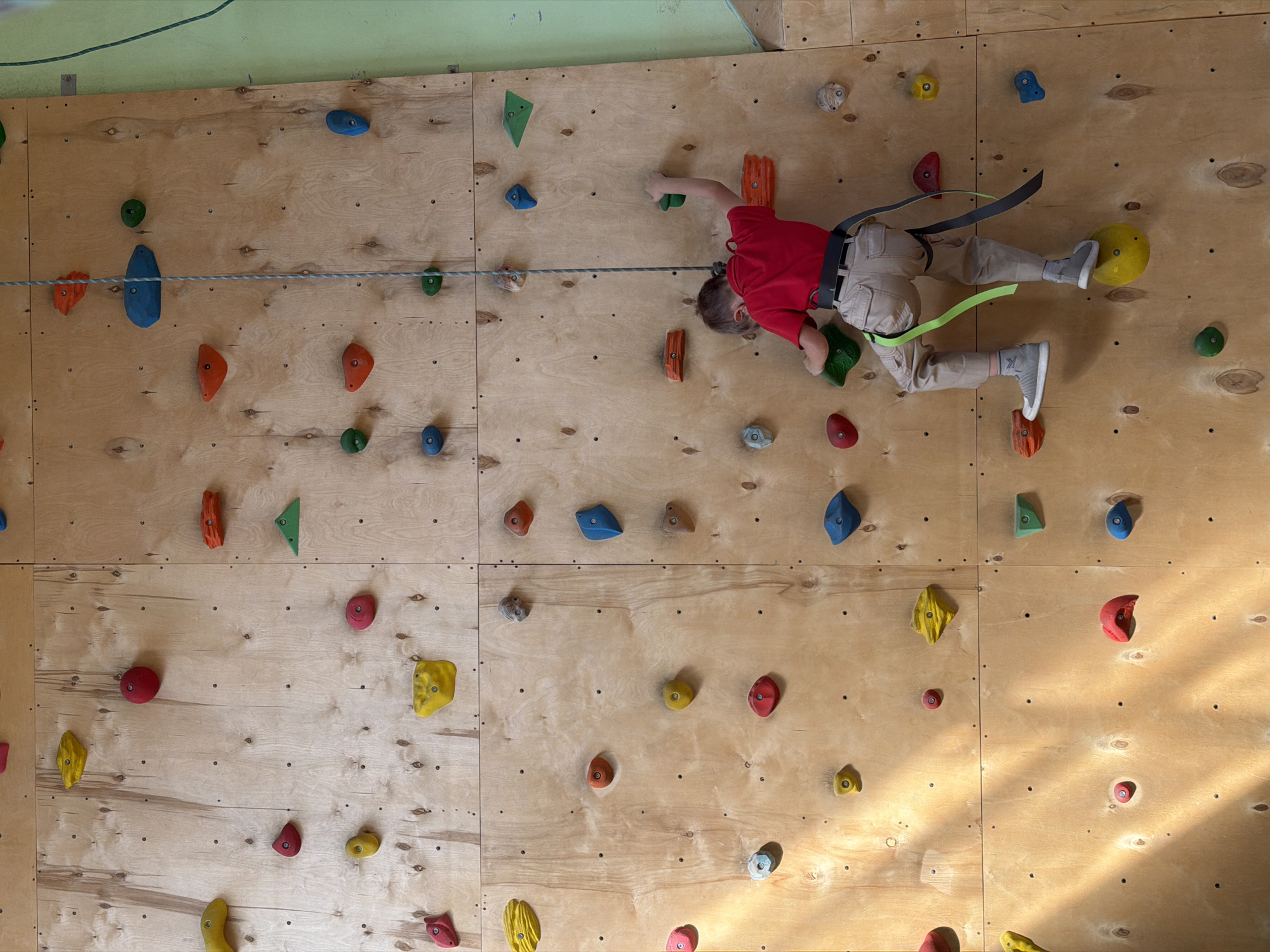 A child is climbing on an indoor rock climbing wall. The wall is made of plywood panels and has numerous colorful climbing holds attached. The child is wearing a red shirt, tan pants, gray shoes, and a climbing harness with green and gray straps. A gray rope is connected to the harness and runs horizontally across the wall. The child is positioned roughly in the center of the frame, with their body oriented horizontally against the wall. Their left hand is gripping a green hold, and their right hand is reaching for an orange hold. Both feet are placed on holds: the right foot is on a yellow hold, and the left foot is on a red hold. The wall appears to be indoors and well-lit. The background shows the top of the wall and ceiling, which are painted light green. The location, Izhevsk, Russia, is not discernible from the visual information.