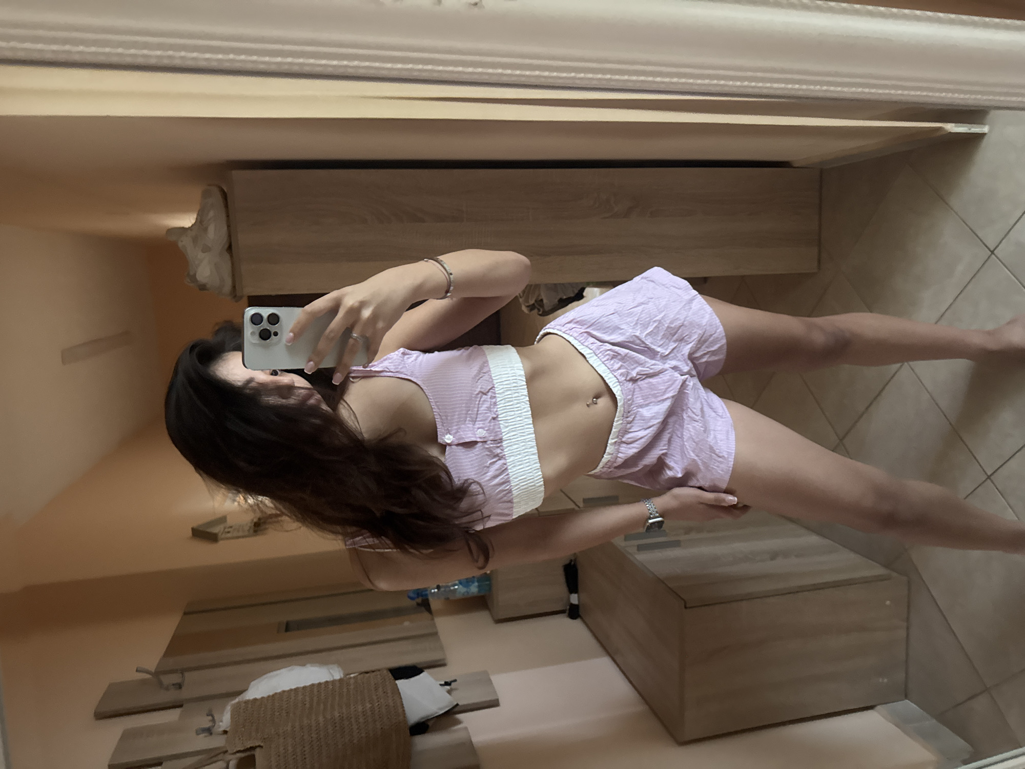 A young woman with dark hair takes a mirror selfie in a hallway, wearing a pink and white striped pajama set consisting of a crop top and shorts. She is holding a smartphone in her left hand, capturing the reflection. The hallway has peach-colored walls and tiled flooring. To the left, there is a wooden dresser with items on it, and a straw bag hanging on the wall. To the right, a tall wooden wardrobe is visible. The overall impression is of a casual, indoor setting, possibly a bedroom or dressing area, located in Plovdiv, Bulgaria.