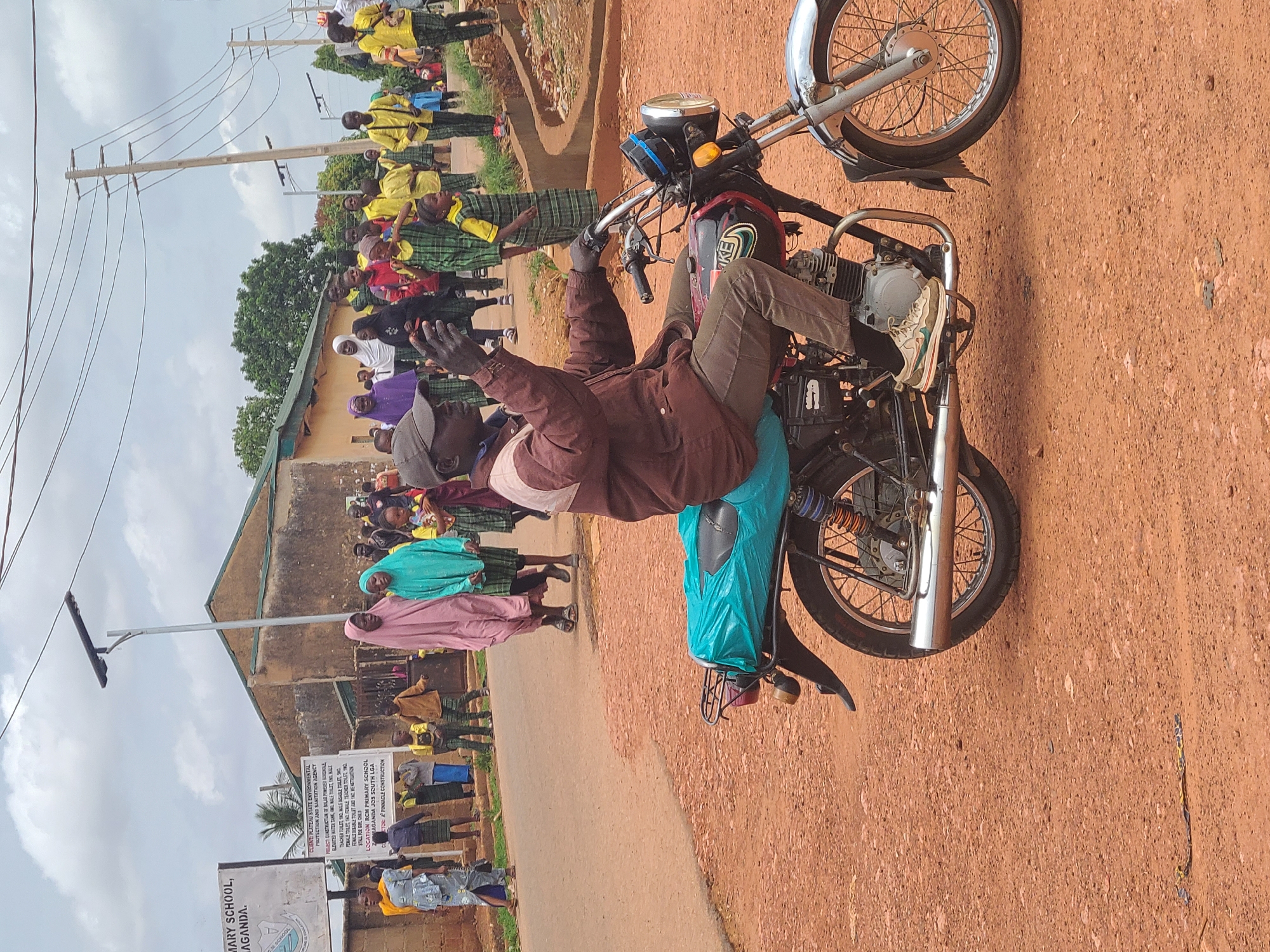 A man, wearing a brown jacket, tan pants, and white sneakers, sits on a motorcycle in front of a crowd of children and adults. The crowd is gathered near a sign indicating "RCM Primary School, Maganda, Jos South LGA" in Jos, Nigeria. The school has a light-colored building visible in the background, and electrical poles line the street. The ground is reddish-brown. The man on the motorcycle has his arms raised. Many people are wearing what appear to be school uniforms consisting of yellow shirts and plaid skirts or pants. Some adults are wearing different colors, including purple, light pink, and turquoise.