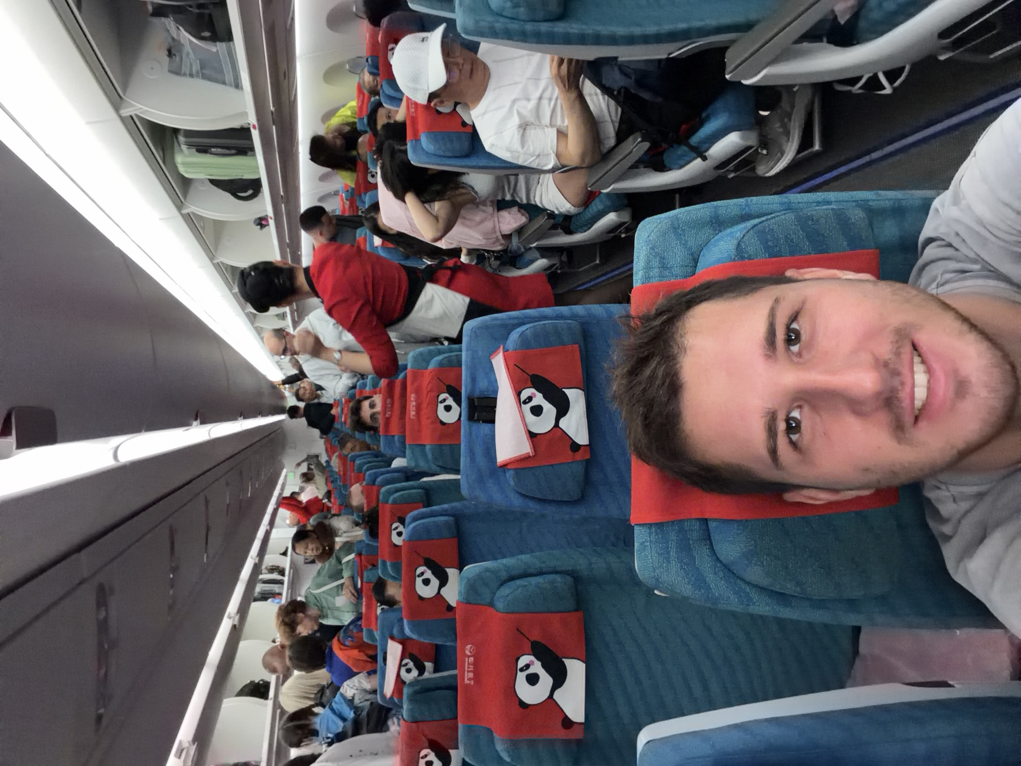 An eye-level selfie captures the interior of a commercial airplane cabin, viewed from the perspective of a male passenger. The image is oriented vertically, showing the length of the aircraft aisle.
In the foreground, the young man taking the selfie is smiling broadly, looking directly at the camera. He has short brown hair, a slight beard, and is wearing a light grey t-shirt. His head rests against a red headrest cover.
Behind him, extending down the aisle, are numerous rows of blue fabric airplane seats. Each seat is equipped with a distinctive red headrest cover featuring a white panda holding a green stalk, possibly bamboo. Faint Chinese characters are visible below the panda design on some of these covers. Small, neatly folded red towels or napkins are also placed on some headrests.
Several other passengers are visible, seated in their respective rows. Some are looking forward, while others are resting or engaged in personal activities. Notable passengers include a man in a white cap and white striped t-shirt further down the aisle, and another wearing a red top closer to the foreground.
Above the seats, the overhead bins are closed, though one is partially open, revealing a light green suitcase and other luggage. The cabin lighting is artificial, illuminating the interior uniformly. The overall scene suggests a routine flight experience, with passengers settled in their seats. The panda motif and Chinese characters on the headrest covers are distinct cultural cues. The plane is within the context of Khimki, Russia, suggesting a flight either originating from, destined for, or transiting through the region.
