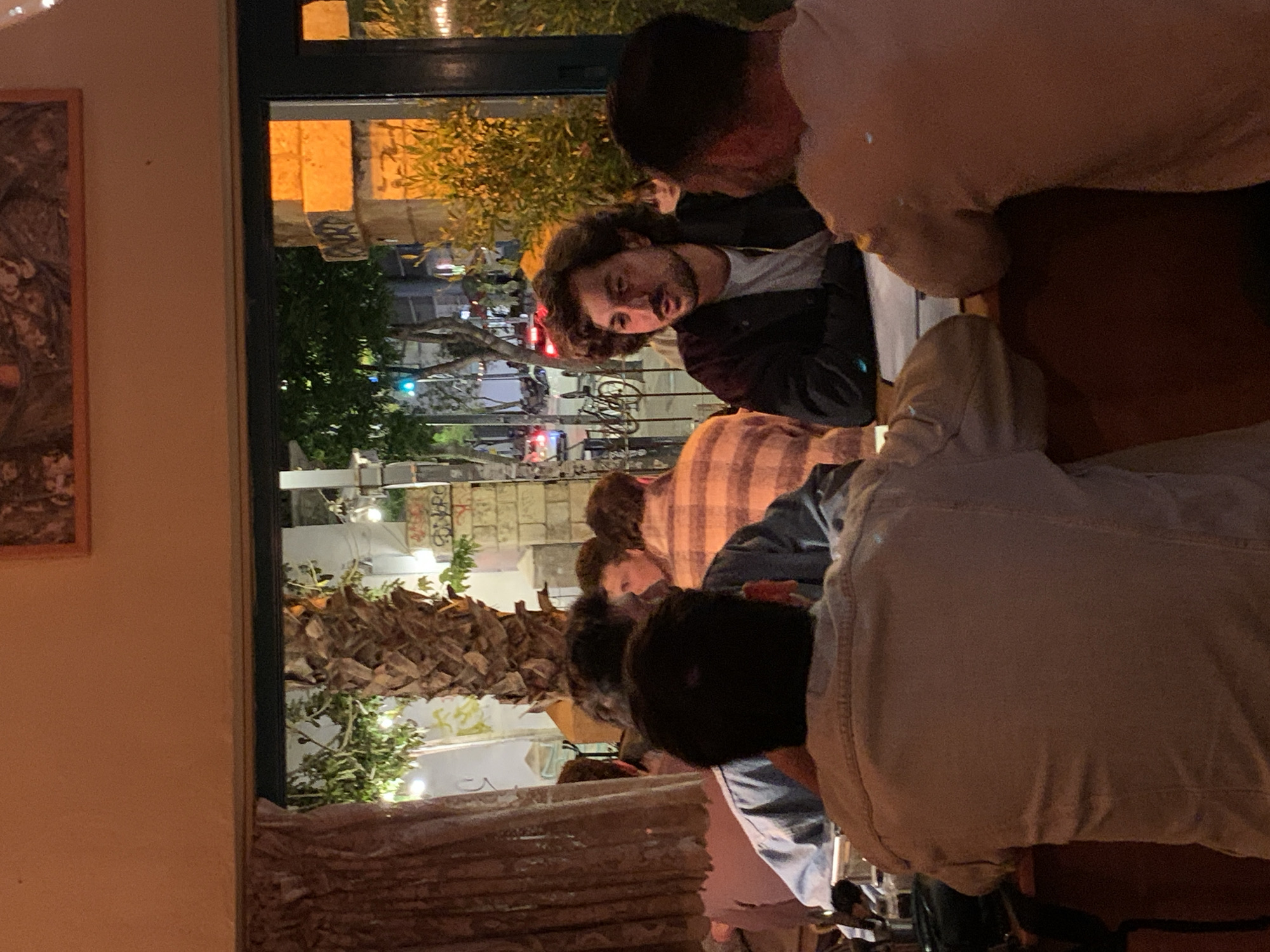 The image displays an interior view of a restaurant or bar located in Tel-Aviv, Israel, at night. The perspective is through a large window overlooking an urban street. Inside, several individuals, primarily men, are seated at tables. In the mid-ground, a man with dark, curly hair, a beard, and a mustache, wearing a dark jacket over a light t-shirt, is prominently featured, facing right and engaged in conversation with another individual whose back is partially visible. Papers or menus are on the table in front of them. Other patrons are seated behind and to the left, partially obscured; one is wearing a plaid shirt and another a light blue shirt. The interior includes brown patterned curtains on the left and a framed picture on the wall above the window. Outside the window, a street scene is illuminated by artificial light. A large palm-like tree trunk stands prominently. Further along the street, leafy trees, streetlights, a green traffic signal, vehicles (with illuminated lights), and parked bicycles are visible. A building exterior to the right of the window displays graffiti, including partial text such as "AKR" and "CONRE."