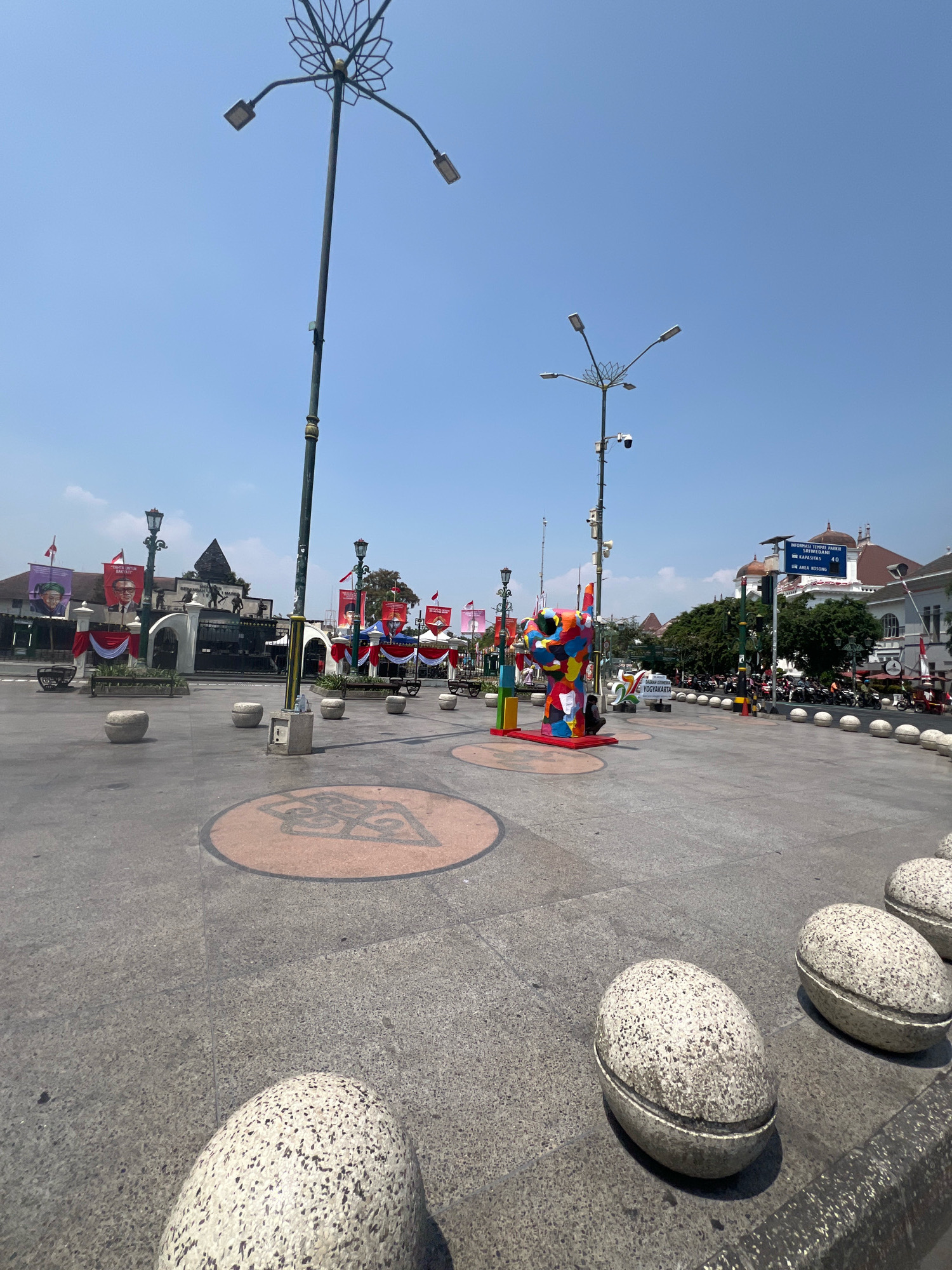 The image captures a wide-angle view of a public square in Yogyakarta, Indonesia, under a clear blue sky. In the foreground, large, egg-shaped concrete bollards are spaced out on the paved ground. In the mid-ground, a vibrant, multi-colored abstract sculpture is prominently displayed on a circular platform. Further back, a tall lamppost with multiple lights stands to the left, and another lamppost with a camera attached is visible on the right.
Beyond the sculpture, a memorial monument with statues is partially visible, flanked by banners and red and white drapes, suggesting a possible event or celebration. Several other lampposts and decorative elements are scattered throughout the plaza. In the background, traditional Indonesian architecture with red-tiled roofs can be seen, along with trees, more banners, and a street scene with parked motorcycles and a few moving vehicles. The overall atmosphere appears to be that of a bustling public space on a sunny day.