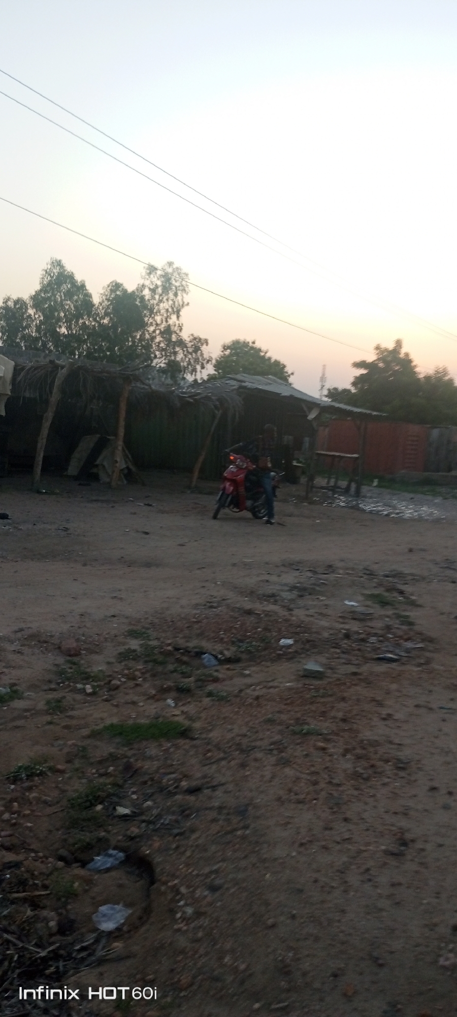 A daytime shot in what appears to be a roadside setting in Lokoja, Nigeria.
**Subjects/Objects:**
* A dirt road or path makes up the foreground, showing some minor depressions, small rocks, and scant vegetation.
* A structure with a thatched roof, supported by wooden posts, stands in the middle ground. Adjacent is a metal or corrugated iron shelter with an attached table.
* An individual stands next to a red motorcycle parked near the thatched-roof structure.
* Trees are visible in the background.
* Power lines stretch across the sky.
**Environment:**
* The sky is bright, suggesting possible dusk or dawn lighting.
**Actions/Interactions:**
* An individual stands beside a red motorcycle.