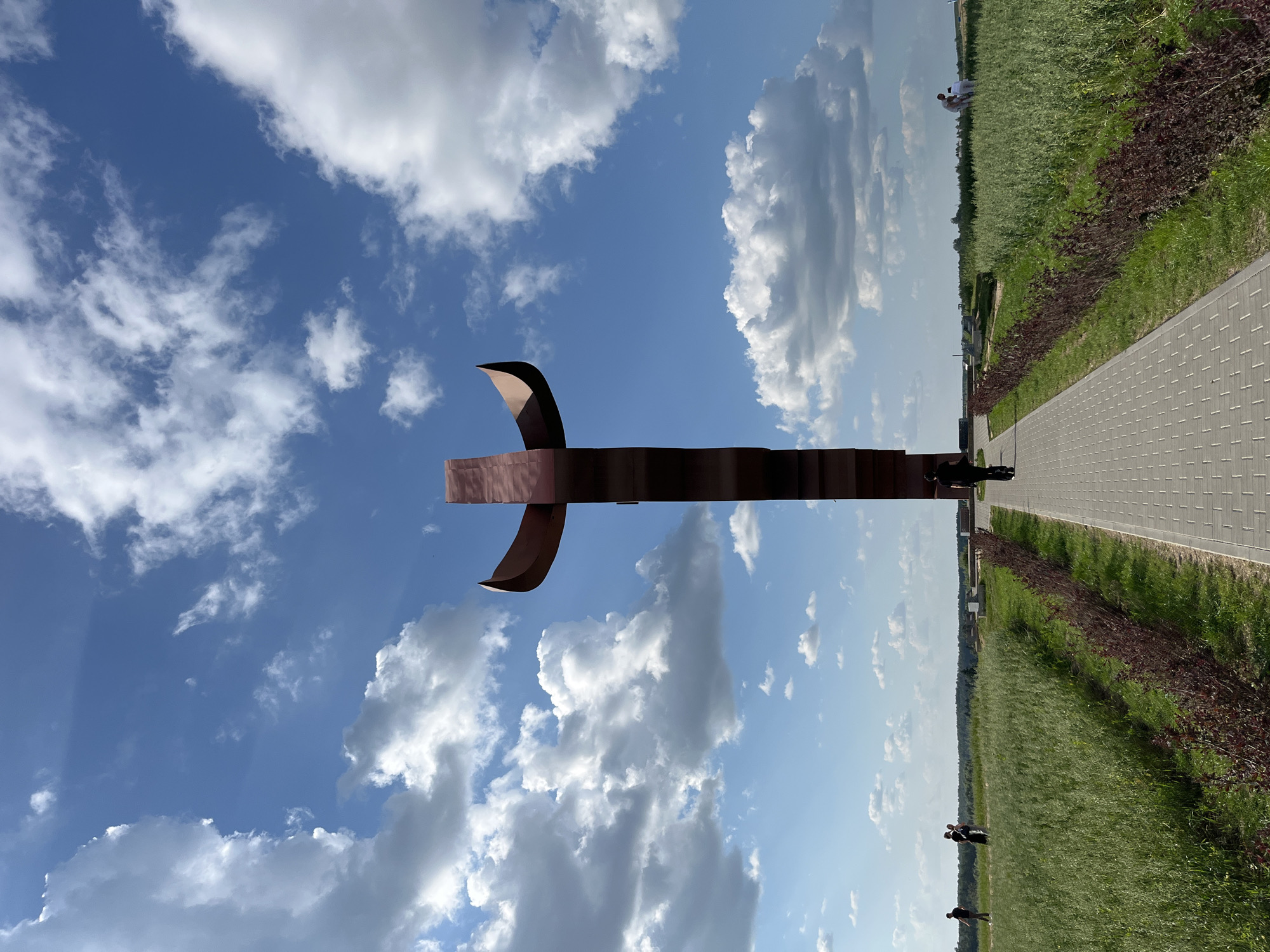 A tall, metallic, cross-shaped monument stands in the center of a paved walkway. The monument is made of reddish-brown material and has curved, wing-like extensions at the top. The walkway leads through a grassy field that stretches to the horizon, where a line of trees is visible. The sky is blue with scattered white clouds. Several people are visible in the distance, walking on the walkway and in the field. The monument is located in the Крошынскі сельскі Савет of Belarus.