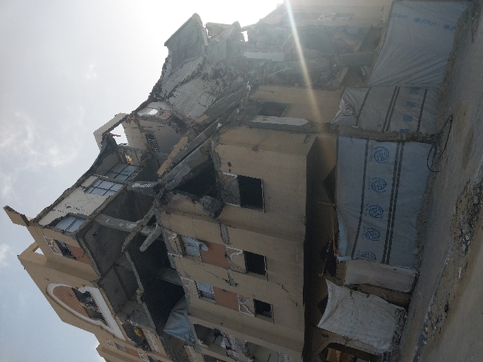 The image displays a severely damaged multi-story building, viewed from a low-angle perspective under bright daylight. The structure shows extensive destruction, particularly on its upper floors, where large sections of concrete walls and slabs have collapsed, exposing twisted rebar and rubble. Remnants of window frames are visible in some sections, but most windows are shattered or missing.
The building's exterior is a light brown or beige color. On the right side of the structure, several sections are covered with white or light blue tarpaulins or sheets. These coverings feature repeating circular logos, clearly identifiable as the UNICEF emblem, with the stylized text "unicef" inside a circle. Below the main UNICEF logo, there is additional, indistinct text or a pattern repeating across the sheets. The presence of UNICEF materials suggests humanitarian aid or temporary measures in response to the damage.
The scene conveys a sense of devastation, likely the aftermath of a destructive event such as conflict or a major disaster. No individuals are visible in the image. The ground adjacent to the building appears to be rough and debris-strewn. The clear sky and bright glare indicate it is daytime. The destruction is consistent with conditions reported in areas like Khan Yunis, Palestinian Territory.
