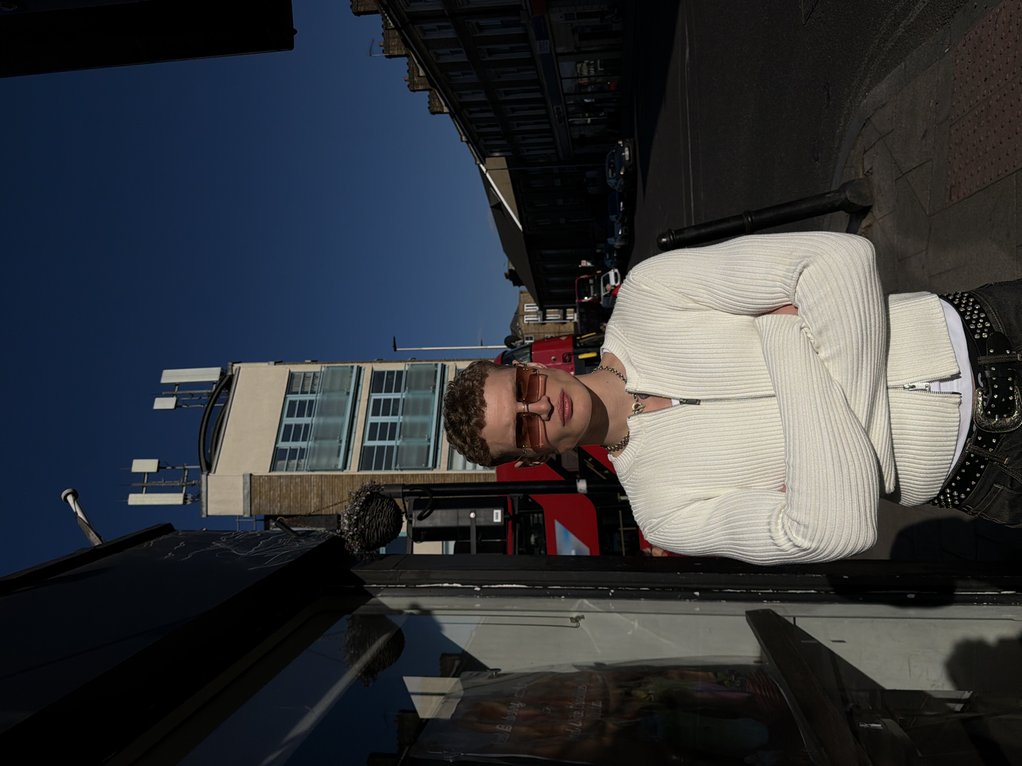 A vertically oriented, ground-level shot captures a young man standing on a city street, with the image rotated counterclockwise. The man, clad in a cream-colored ribbed sweater, dark pants with a studded belt, and sunglasses, is positioned toward the right of the frame with his arms crossed. He is facing the camera.
Behind the man, a London bus is partially visible, followed by other vehicles parked along the curb. Tall buildings rise in the background. The architecture suggests an urban setting, characteristic of London, United Kingdom. The photograph's composition includes reflections and shadows, which add depth to the scene. The background includes a light blue sky.