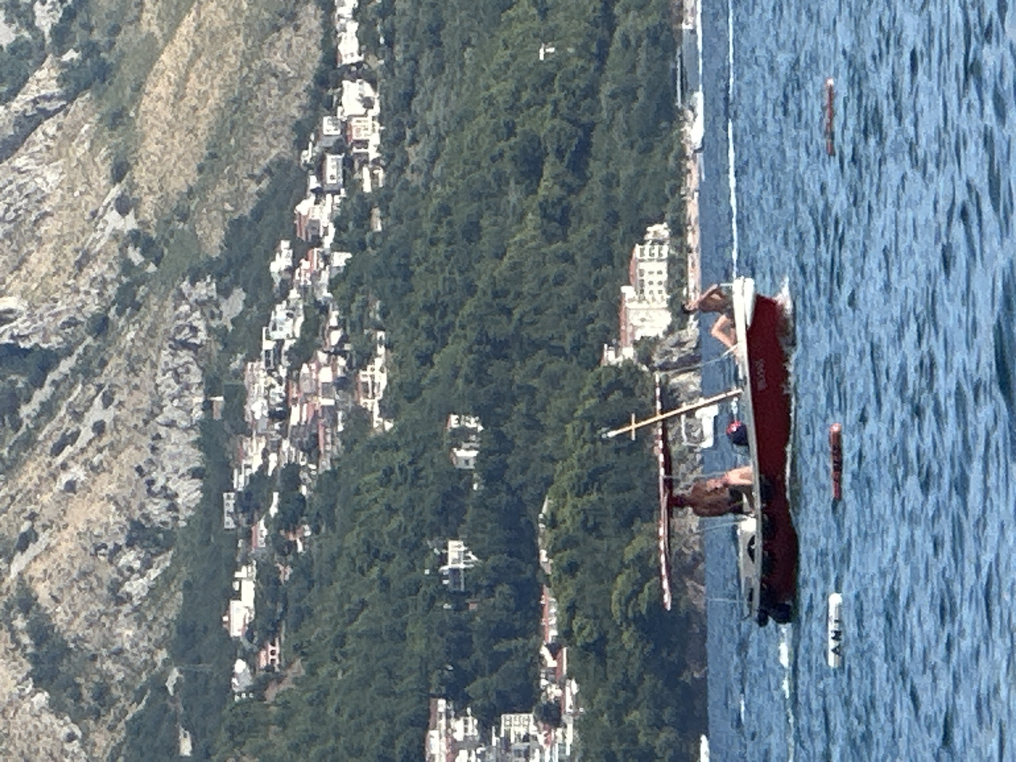 An eye-level, medium shot captures a red motorboat with two people navigating the blue, choppy sea. A person wearing dark shorts stands at the rear of the boat, facing away from the camera and towards the stern. To their right, a second person, also wearing dark shorts, is seated on the edge of the boat, their back to the camera. The boat has a tan awning extending over its midpoint. In the foreground, two orange-red buoys float on the water, one appearing cylindrical and the other elongated. The background depicts a densely forested green mountainside dotted with white buildings, indicative of a coastal town. The upper portion of the image reveals a rocky, barren mountain range above the treeline. The scene is set in Boreti, Montenegro.