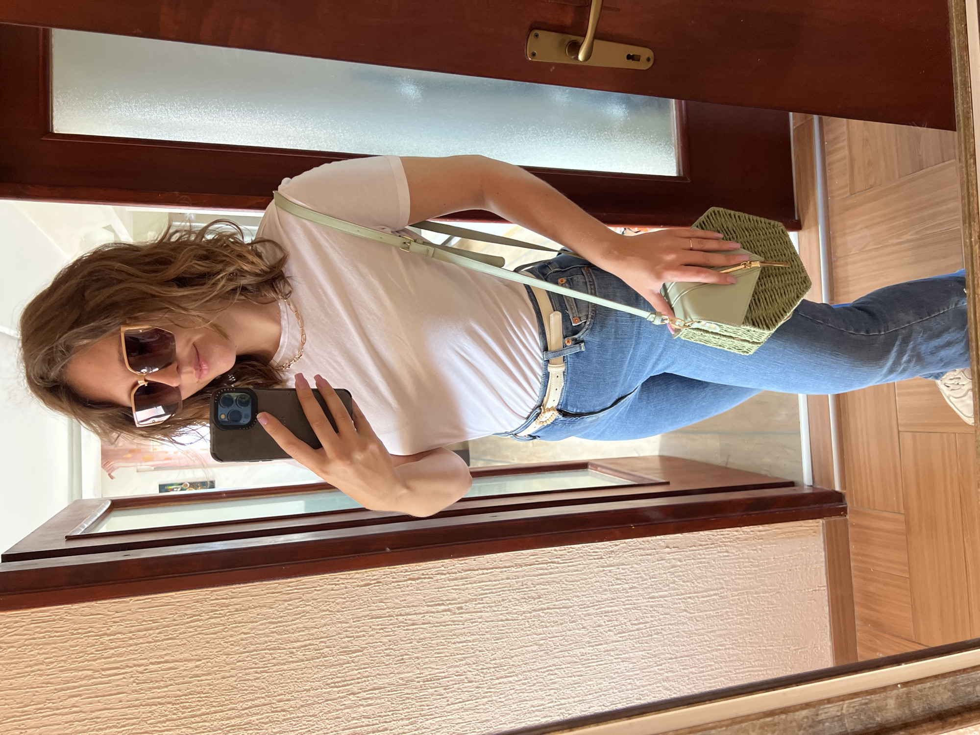 A mirror selfie captures a young woman standing indoors. She has medium-length wavy brown hair and is wearing a white short-sleeved t-shirt, light blue jeans, a light-colored belt, and partially visible light-colored shoes. She accessorizes with large brown sunglasses with gold frames, a gold-colored chain necklace, and light pink nail polish.
In her left hand, she holds a dark-colored smartphone, likely an iPhone, which she uses to take the photo. Her right hand holds a distinctive hexagonal bag made of woven straw or rattan in a muted green shade, featuring a light green flap and strap.
The background shows a dark brown wooden door with a brass-colored handle and lock, and a frosted horizontal glass panel. A light-colored, textured wall is visible to the lower left, and a light brown wooden floor with a parquet pattern extends to the lower right. The overall impression is a bright, daytime indoor setting. There is no visible text in the image. The provided context indicates the location is Podgorica, Montenegro, though the image itself does not contain specific visual cues to confirm this.