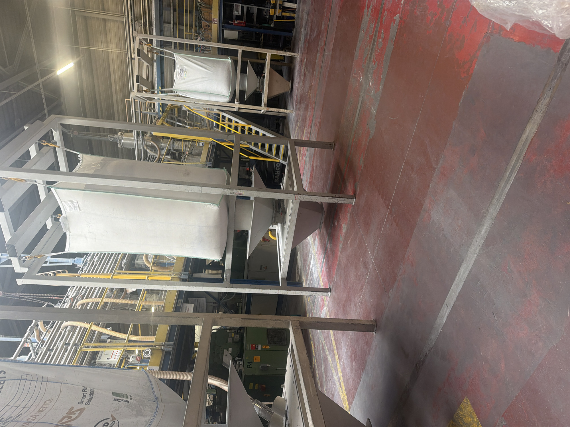 The image depicts an industrial setting with large white fabric sacks, commonly known as bulk bags or FIBCs (Flexible Intermediate Bulk Containers), suspended from metal racks. These sacks appear to be filled with a white powdery substance, likely industrial material.
The environment is characterized by heavy machinery, including piping systems, control panels, and industrial equipment, suggesting a manufacturing or processing facility. Yellow railings and staircases are visible, indicating an elevated work platform or access point. The flooring is a red industrial surface, showing signs of wear and tear with some grey markings and paint peeling. The lighting appears to be from overhead industrial lamps.
There are no people clearly visible in the image. The focus is on the large sacks and the surrounding industrial infrastructure. The overall scene conveys a sense of an operational factory floor.
Regarding the location of Jasin, Poland, there is no explicit visual information in the image that can confirm or deny this specific location. The image shows a generic industrial interior that could be found in many manufacturing plants worldwide.