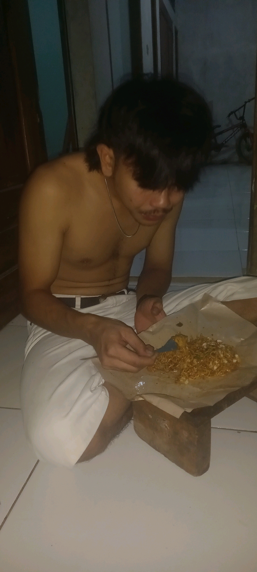 The image shows a young man sitting on the floor and eating from a piece of paper. He is shirtless, wearing white pants and a black belt. He has dark hair, a mustache, and is wearing a thin chain necklace. The man is holding a fork in his right hand and is about to eat some noodles from the paper. The noodles are in a pile on the paper, which is resting on a wooden object. The scene appears to be indoors, with a tiled floor and a dark background that includes a bicycle. The lighting suggests it might be evening or nighttime. The man's expression is neutral as he focuses on his meal. The image was taken in Argasoka, Indonesia.