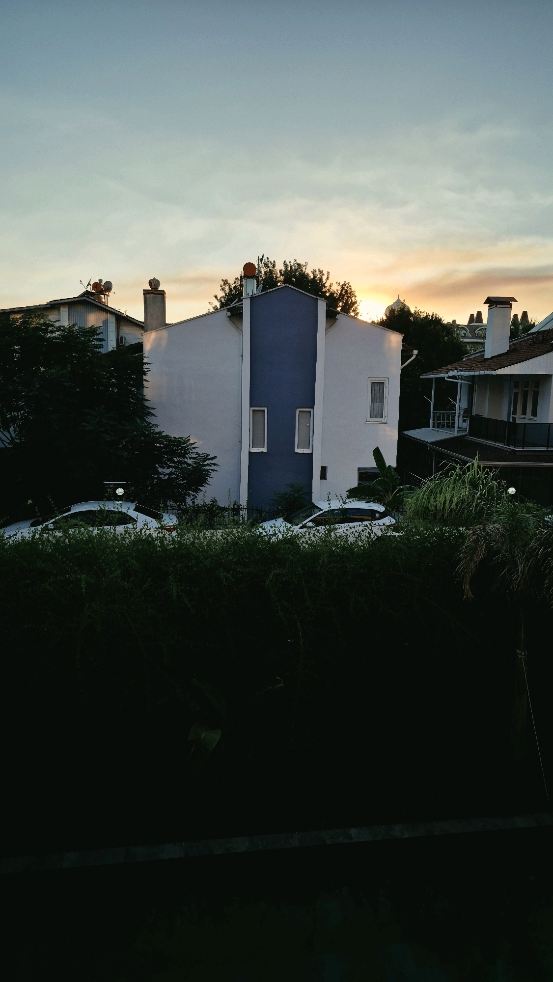 A view of a residential street in Serik, Turkey, at sunset. The sky is a blend of pale blue and soft orange hues, with wispy clouds. In the foreground, lush green foliage obscures the lower portion of the scene, with a dark, horizontal ledge or barrier visible at the very bottom.
Behind the foliage, two white cars are parked side-by-side, their shapes partially visible. A dark metal fence runs behind the cars, separating them from the buildings.
Dominating the mid-ground are two houses. The house on the left is primarily white, with a grey chimney topped with an orange cap. To its right, the main house in the image is a two-story structure. Its facade is mostly white, contrasted by a prominent vertical section of deep blue in the center. This blue section features two narrow, vertical rectangular windows. To the right of this blue section, a single, larger rectangular window is visible on the white wall.
To the right of the blue-and-white house, another house is partially visible. It has a white exterior, a dark roof, and a white balcony with a railing. A chimney with a rectangular cap is also visible on this house. Trees with dark green foliage are scattered throughout the scene, some taller than the houses. The overall impression is of a quiet residential area during the golden hour.