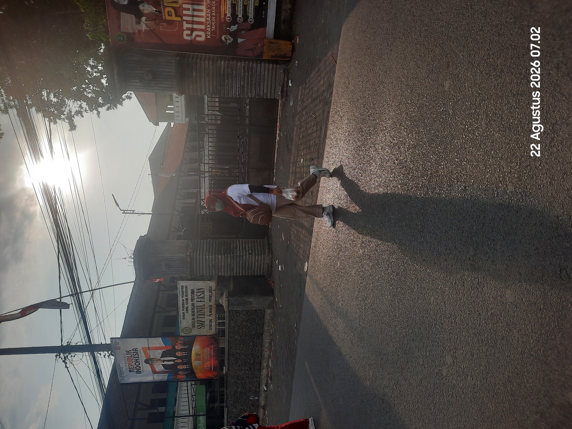 A person wearing a hijab and a mask walks on a street in Pajarakan Kulon, Indonesia, during the day. The sun is low in the sky, casting long shadows. The person is carrying a backpack and a plastic bag. In the background, there are buildings, including what appears to be a school, with banners displayed. The street is paved with asphalt and there is some debris scattered on the side. A date stamp indicates "22 Agustus 2026 07.02" which translates to "August 22, 2026 07:02".