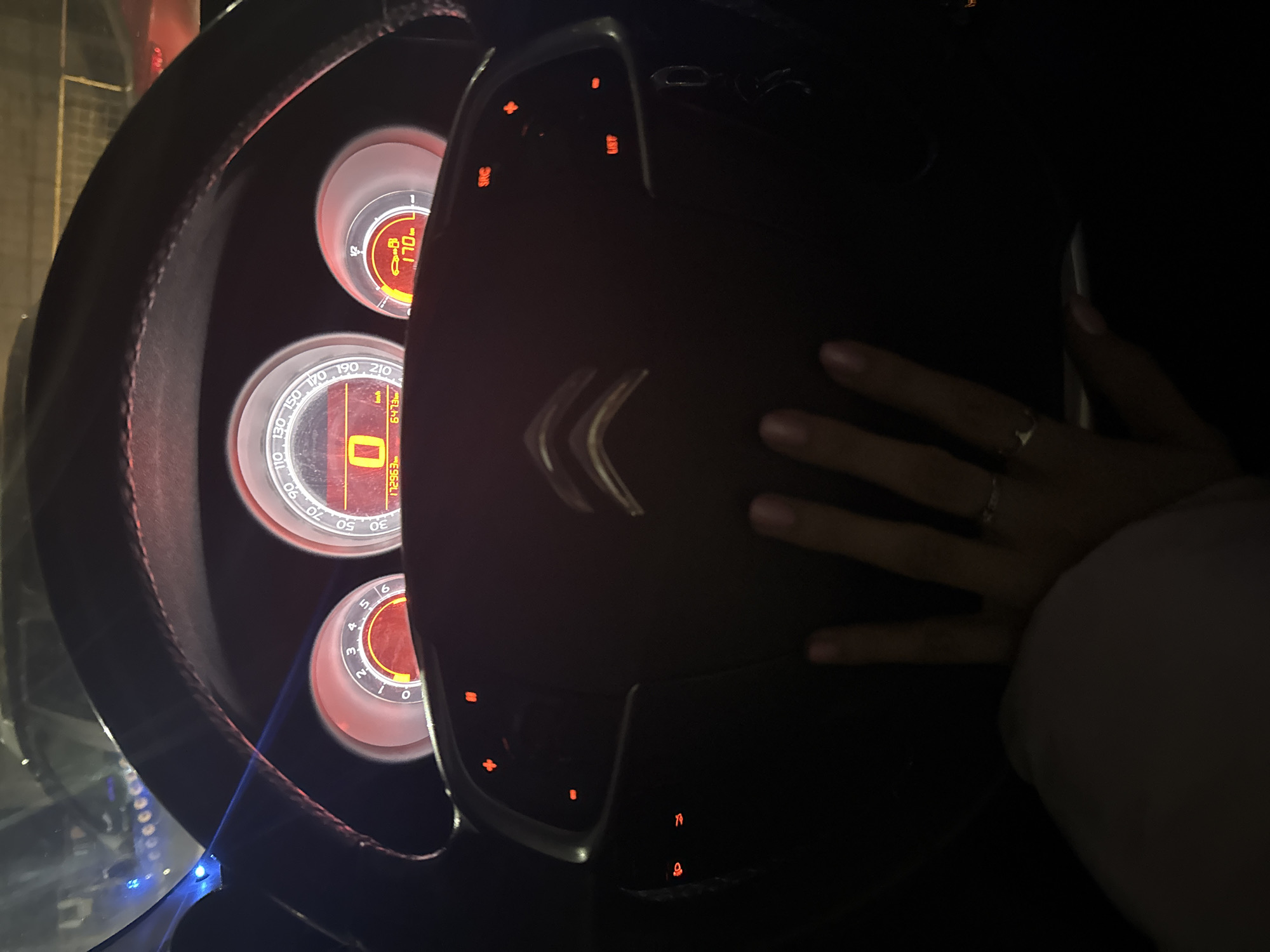 The image displays the dark interior of a car at night, focusing on the steering wheel, instrument cluster, and a person's left hand. The hand, wearing a ring on the ring finger, rests gently on the bottom right portion of the steering wheel. The steering wheel itself is dark and features the distinctive silver Citroën logo at its center. Various control buttons on the steering wheel are illuminated in red, including those labeled "SRC" and "LIST," alongside volume/track controls, phone controls, and other symbols.
The car's instrument cluster, glowing with orange and red light, consists of three circular gauges. The leftmost gauge appears to be an RPM meter, marked from 1 to 6. The central gauge is a speedometer, clearly displaying "0 km/h," indicating the car is stationary. This gauge's digital display also shows "6473" (likely a trip meter) and "172963" (the odometer). The speedometer's scale extends up to 210 km/h. The rightmost gauge shows a "1/2" marking and a digital readout with "170" and "OLI," possibly indicating oil temperature, fuel level, or another vehicle status. The overall darkness of the car's interior is starkly contrasted by the bright, illuminated dashboard and steering wheel controls. Faint blue light reflections are visible on the left side of the frame, suggesting ambient lighting. The scene is set in Izhevsk, Russia.