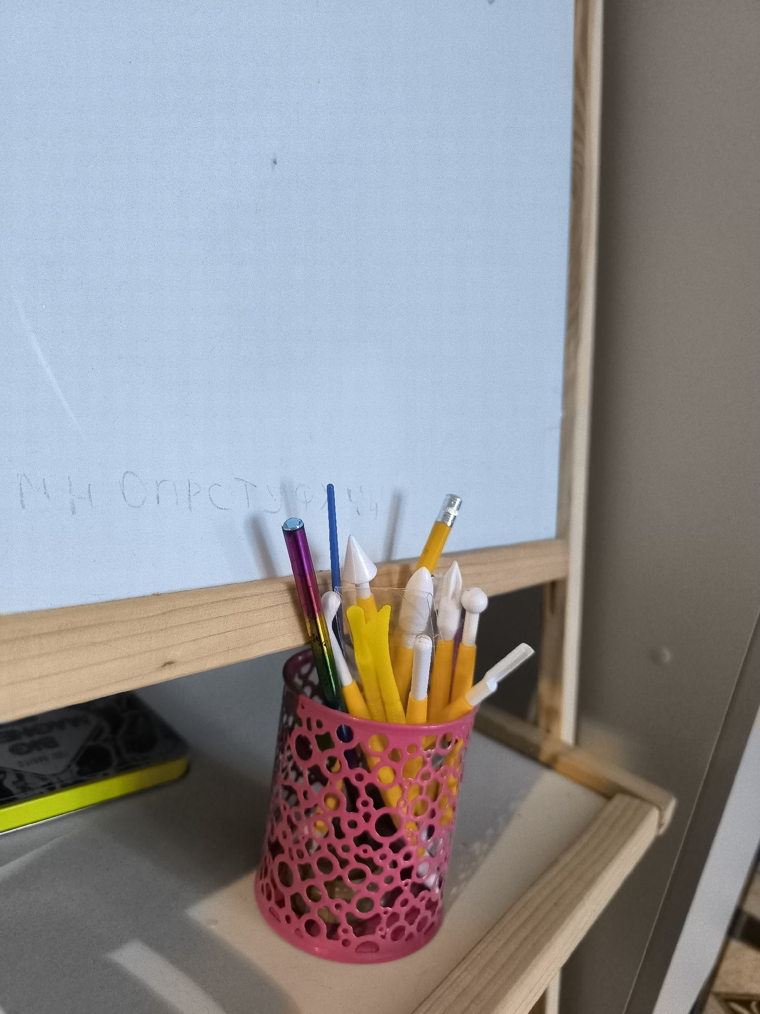 The image shows a close-up of a section of a whiteboard with some faint writing on it, and a collection of art supplies in a pink holder. The writing on the whiteboard appears to be in Cyrillic script. The art supplies include various sculpting or modeling tools with colorful handles, such as yellow, white, and purple, and possibly some pencils. The holder is a pink, decorative mesh container. In the background, a wooden easel is partially visible. A portion of a shelf or desk is in the foreground, with a dark object with a bright yellow edge on it. The overall scene suggests a creative workspace, possibly a child's art area or a craft corner.
There is no information in the image that suggests a specific location within the city of Horishni Plavni, Ukraine, or any other city for that matter. The provided description is limited to the visual elements present in the photograph.