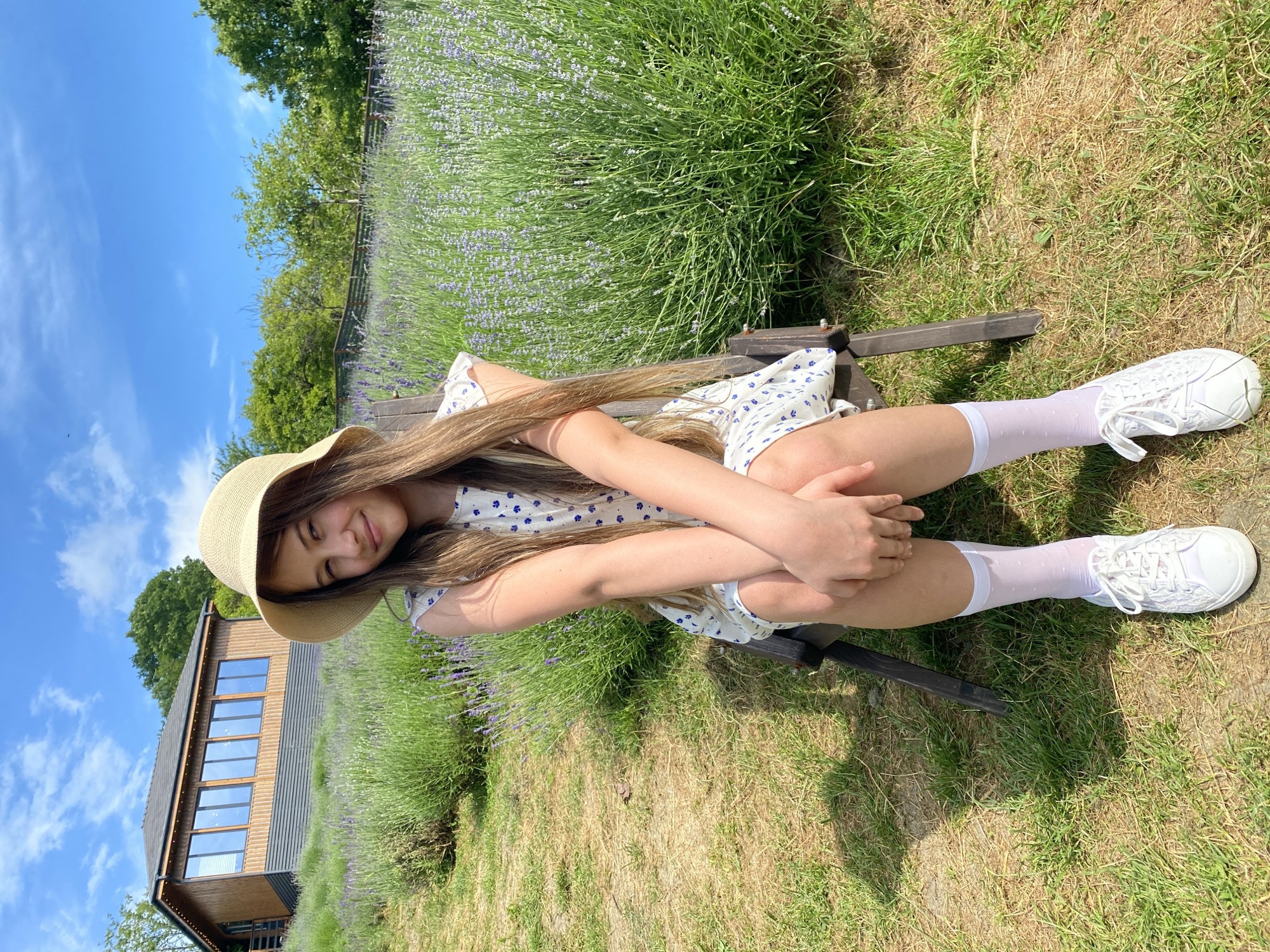 A young girl with long, brown hair, wearing a straw hat, a white floral dress, white socks, and white sneakers, is sitting in a wooden chair in a lavender field. She is looking at the camera and smiling. In the background, there is a wooden building on a hill, trees, and a fence. The sky is blue with white clouds. The location is Verkhneye Uchdere, Russia.