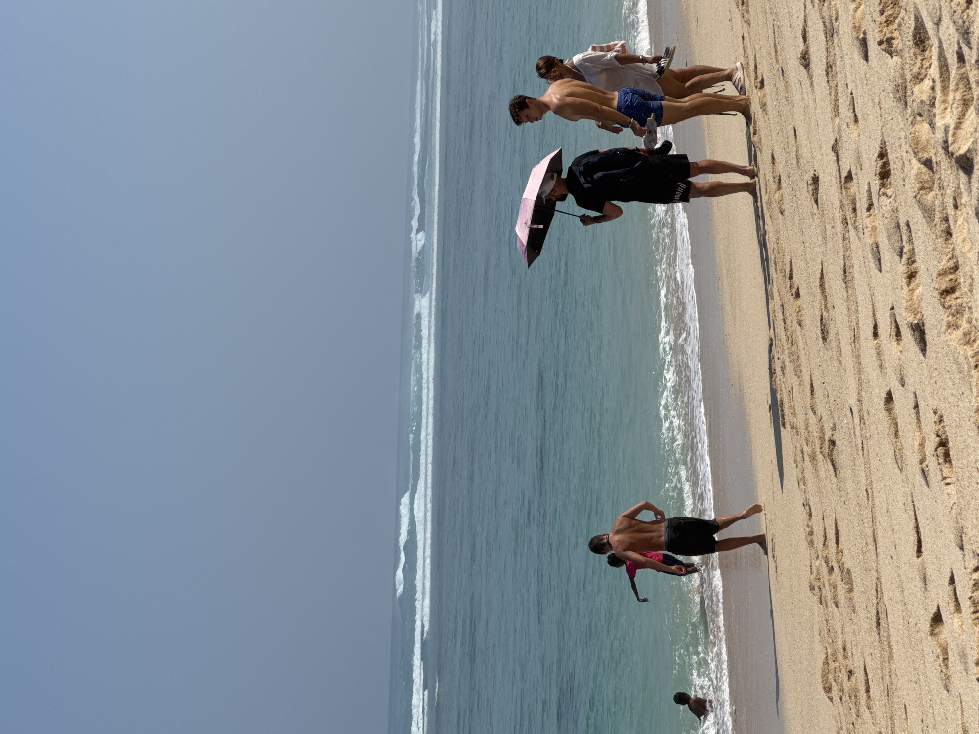 In Kuta Selatan, Indonesia, on a sandy beach, a group of people are present. The ocean features several lines of breaking waves under a hazy sky. In the foreground, the sand is textured with footprints and small mounds. On the beach, four individuals are observed. Two men and a woman are standing closer to the water, facing the ocean. One man is shirtless, wearing blue swim trunks, and holding a shoe. The woman is wearing a white cover-up and holding a shoe. Another man, facing away from the camera and towards the ocean, is holding a pink umbrella over himself. He is wearing a black t-shirt and black shorts, and carrying a black backpack. In the background, two young individuals are in the shallow water. One is wearing a bright pink shirt and black shorts, and the other is mostly submerged in the water. The overall scene depicts a coastal setting with moderate ocean activity and people engaging in beach activities.