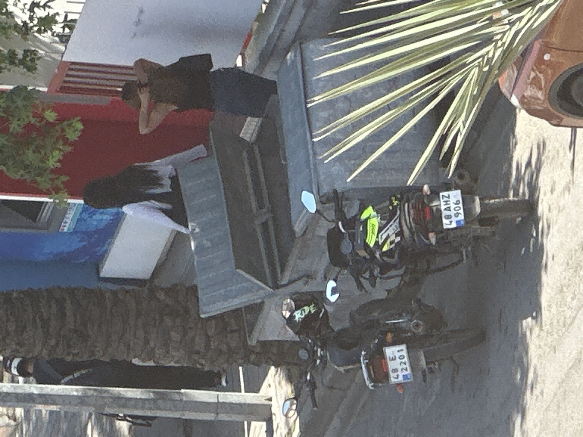 The image captures a street scene in Fethiye, Turkey. Two motorcycles are parked on the right, with license plates visible: one reads "48 AHZ 906", and the other reads "48 E 2201". In the background, two people are seen near buildings and a palm tree. One person is sitting on a ledge or bench, while the other appears to be walking. Buildings with a red and white facade are visible, along with a blue awning. A corrugated metal structure is located near the motorcycles. The scene is lit by natural sunlight.