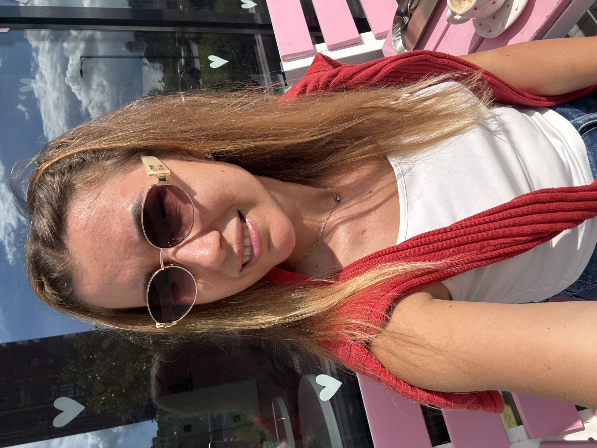 A woman with blonde hair and sunglasses is taking a selfie while sitting on a pink bench outdoors. She is wearing a white tank top and a red sweater draped over her shoulders. A cup of coffee and a plate with hearts are visible on the bench next to her. In the background, there is a building with a reflection of the sky and clouds in its windows, and a street lamp. There are white heart stickers on the window. The scene appears to be in a city, possibly a cafe or restaurant.
