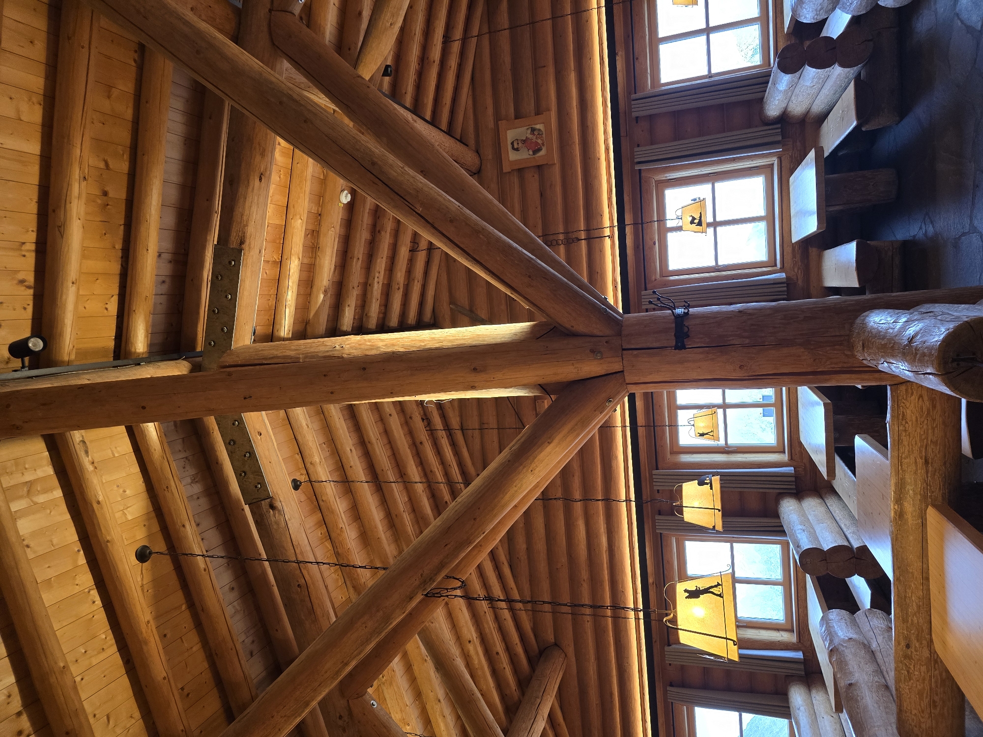 The image shows the interior of a rustic log cabin restaurant. The ceiling is made of exposed wooden beams and planks. Several pendant lights with lampshades depicting hikers are suspended from the ceiling. Large wooden log pillars support the roof structure. The walls are also constructed from logs. In the background, there are rows of wooden tables and benches. Windows with light curtains are visible along the walls, letting in natural light and offering a view of the outside landscape, which appears mountainous. A framed portrait is hung on one of the wooden walls. The floor is made of dark stone tiles. The scene depicts a cozy, traditional dining area designed to evoke a mountain lodge ambiance. The location is identified as Garmisch-Partenkirchen, Germany.