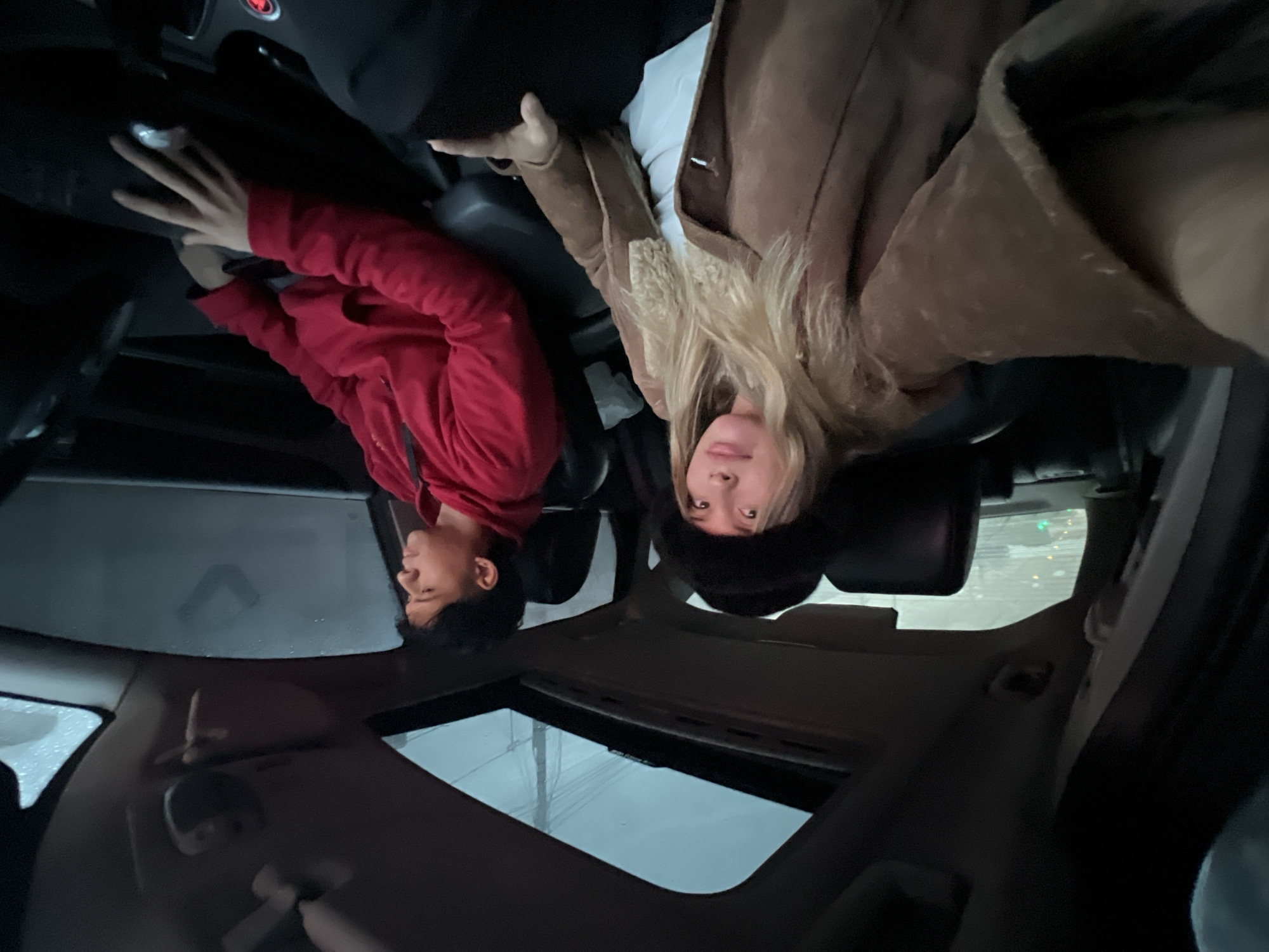 Two individuals are visible inside a vehicle with a sunroof. One person wears a red hoodie and the other a brown jacket with a fur collar. Rain is visible on the sunroof. The location is unknown.