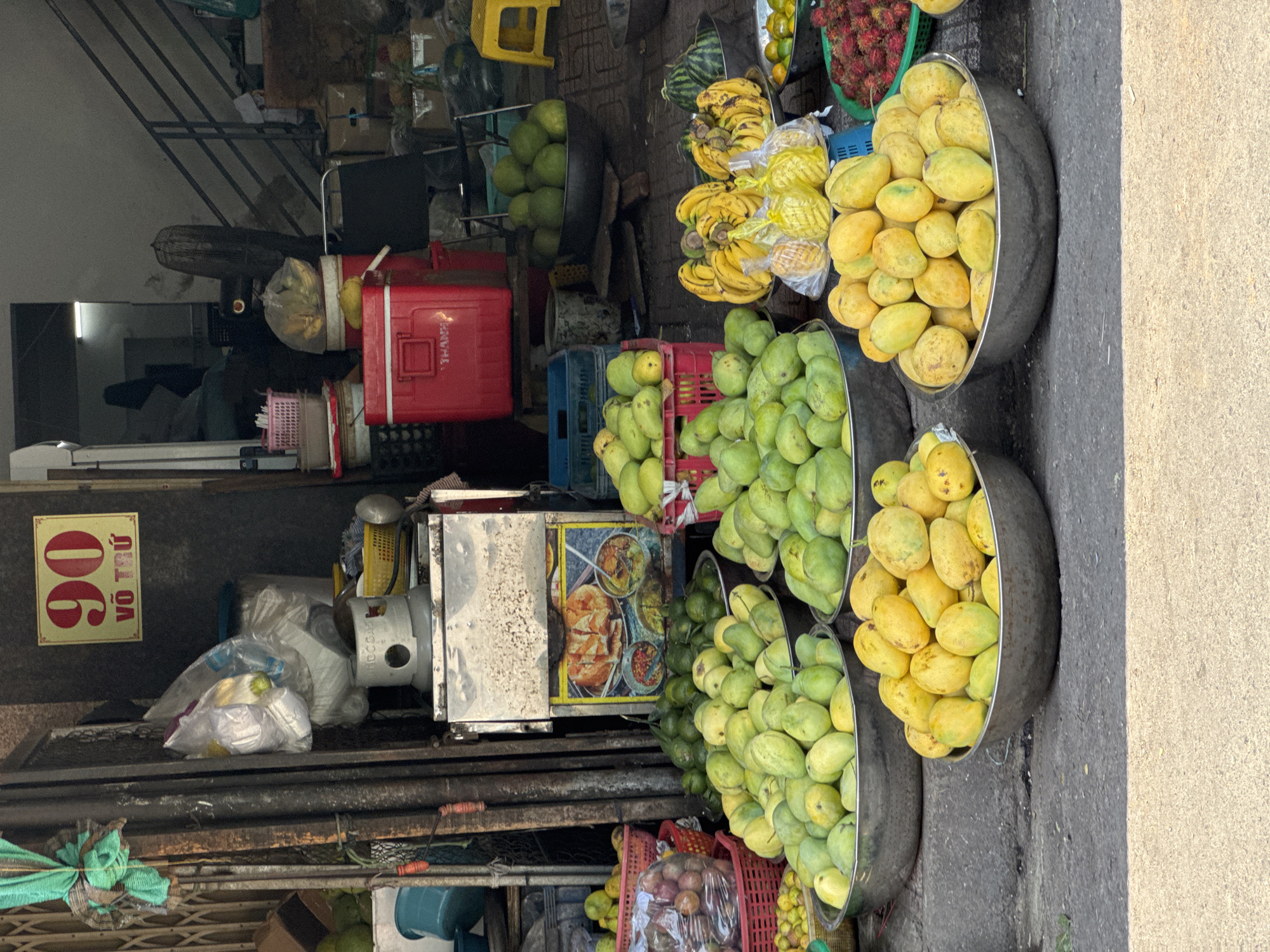 This image depicts a vibrant outdoor fruit market stall in Nha Trang, Vietnam. The stall is filled with a variety of fresh fruits displayed in large metal bowls and red plastic crates. Prominently featured are numerous green and yellow mangoes, as well as bunches of yellow bananas. Other fruits visible include what appear to be lychees or rambutans in a green basket, and green fruits resembling pomelos or large citrus. In the background, a red cooler box and other market-related items are visible, along with a sign displaying the number "90" in red and Vietnamese text. The market appears to be set up on a paved street or sidewalk. No people are clearly visible in the image, but the presence of the market suggests it is an active commercial area. The overall impression is one of abundant fresh produce typical of a bustling Vietnamese market.