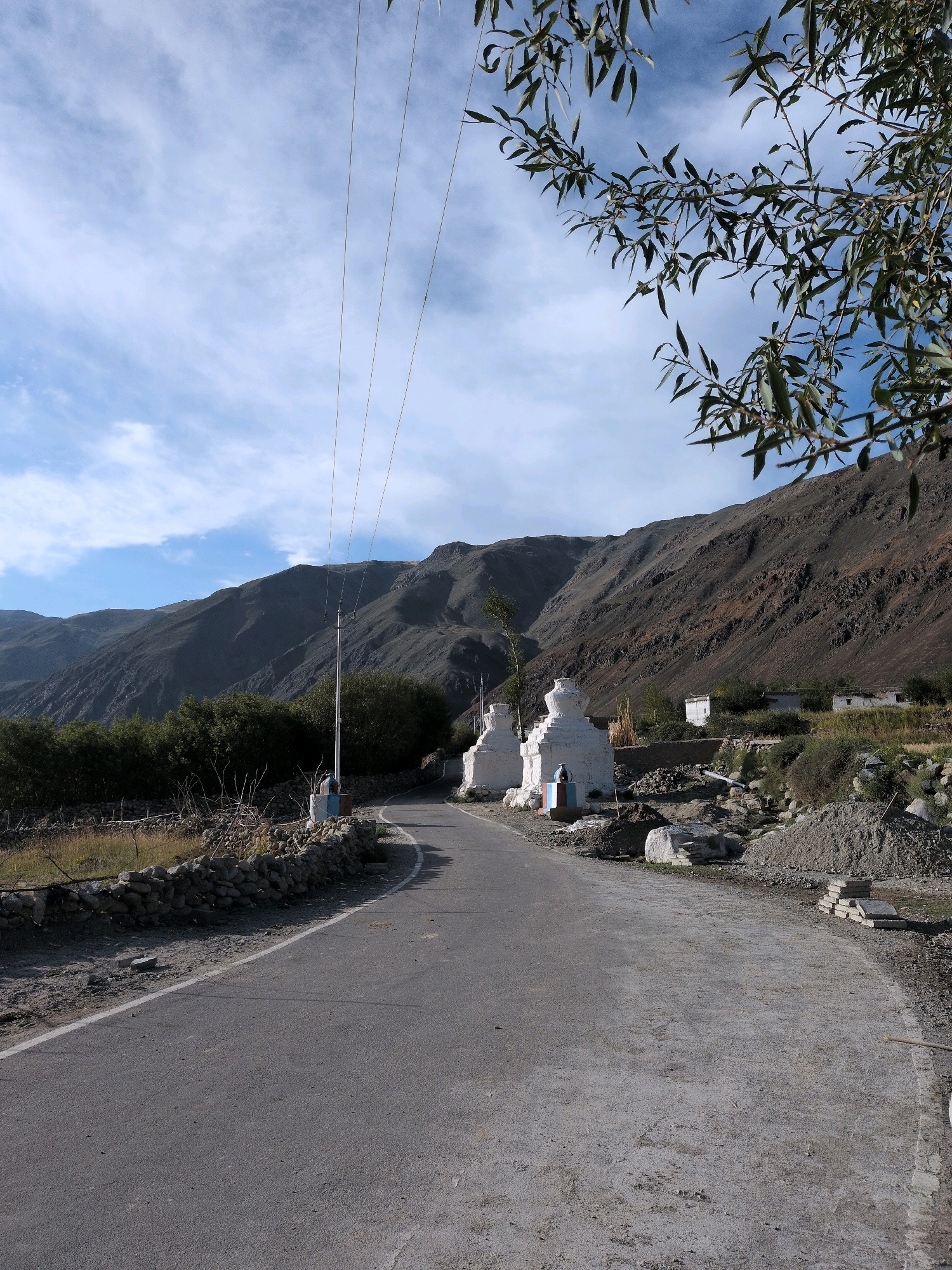 A road winds through a mountainous landscape in Skidmang, India. Two white stupas stand beside the road on the left, and a pile of dirt and rocks is on the right. A stone wall runs along the left side of the road, separating it from a field. Trees and bushes are visible in the middle ground, and steep, barren mountains rise in the background. The sky is blue with scattered clouds. Power lines stretch across the scene.