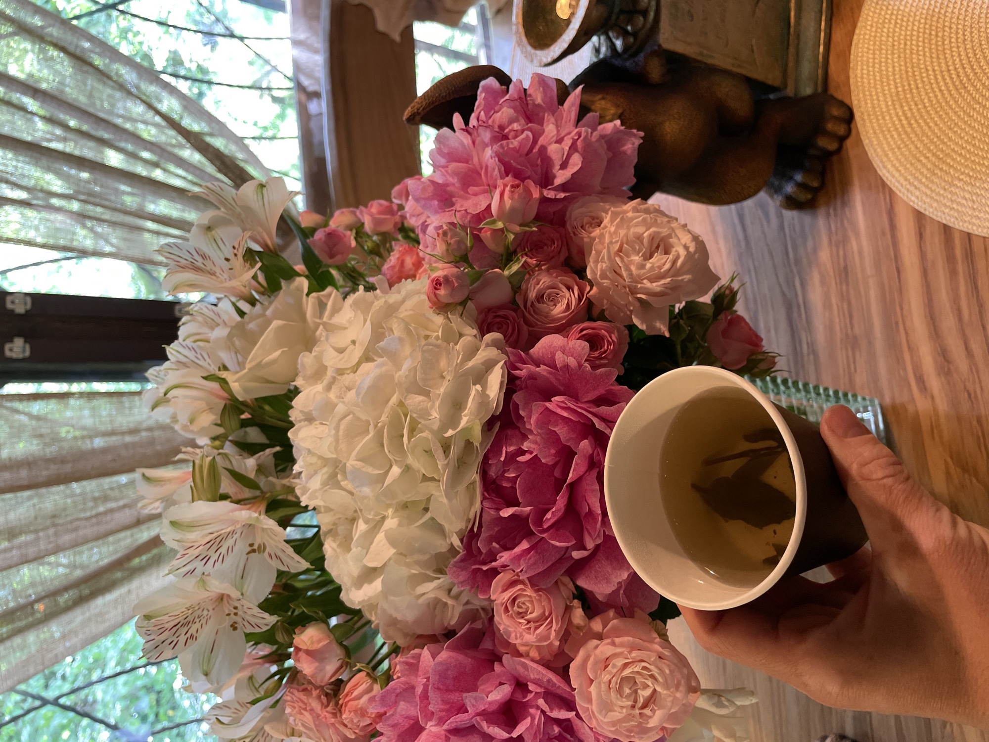 The image shows a close-up, high-angle view of a bouquet of flowers with a cup of tea held in a person's hand, likely located indoors in Solokh-Aul, Russia.
The floral arrangement consists of white hydrangeas, pink peonies, pink and white roses, and alstroemeria with pink stripes. The flowers are densely packed together.
A hand is holding a tea cup with liquid visible inside. The cup is brown on the outside and white on the inside, with tea leaves floating inside.
In the background, out of focus, there's a window with blinds. Also present is a bronze-colored decorative item resembling a cherub or animal figurine. Beside the figurine is a straw hat.
The scene suggests a serene, domestic setting.