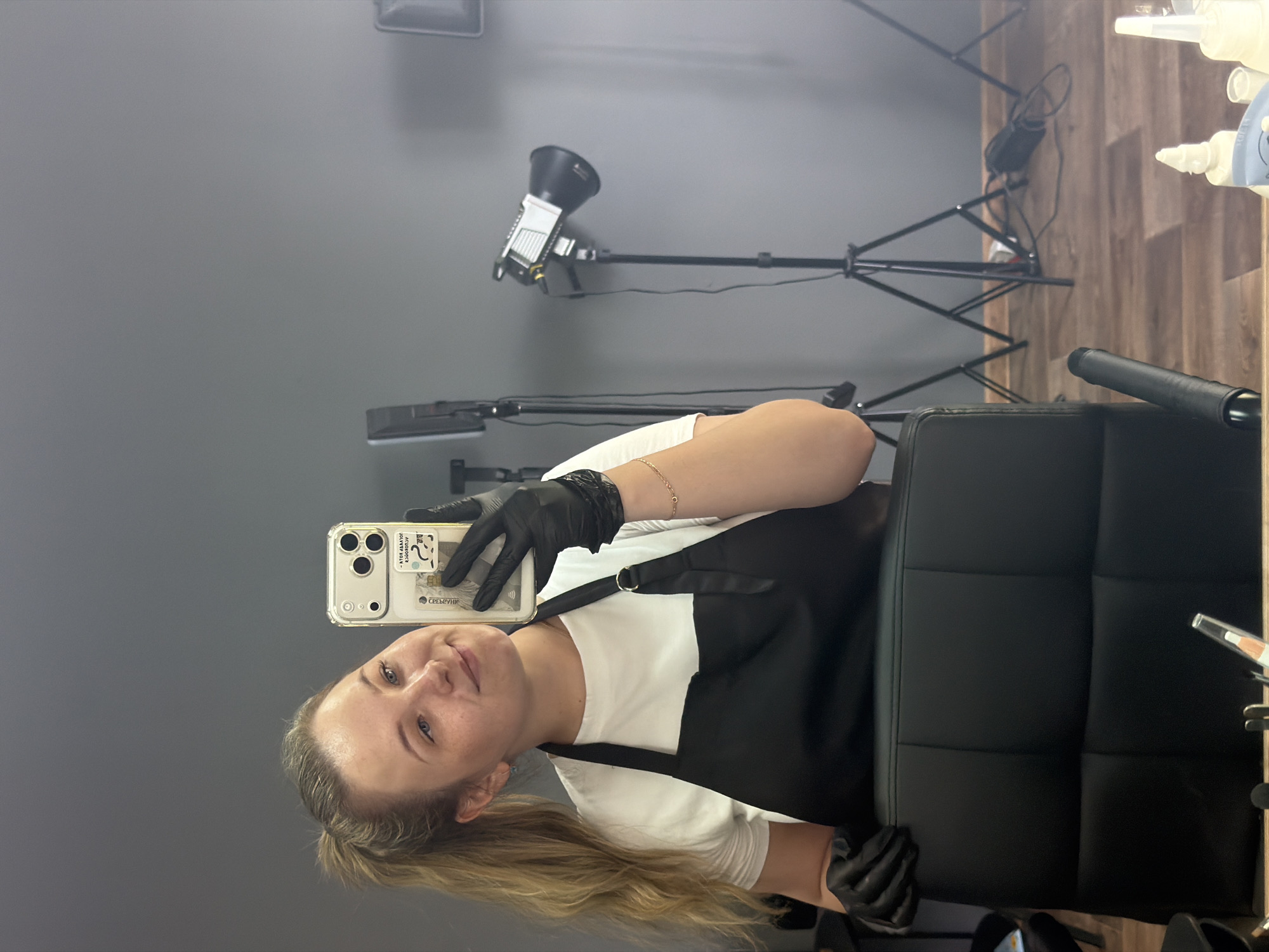 A blond-haired woman is taking a selfie in a salon or studio. She wears a white t-shirt under a black apron and black gloves. She is holding an iPhone in her left hand.
The background is a gray wall with two studio lights on tripods. A black chair is to the right of the woman. Bottles of what appear to be hair styling products are visible near the right edge of the frame on a shelf with wooden paneling.
The location is potentially in the city of Chaikovsky, Russia, as indicated by the text visible on the woman's phone in the image. The text, partially obscured, includes the word "СБЕРБАНК", which translates to "Sberbank" in English, a common Russian bank. This information, combined with the requested city, suggests the image's potential origin.