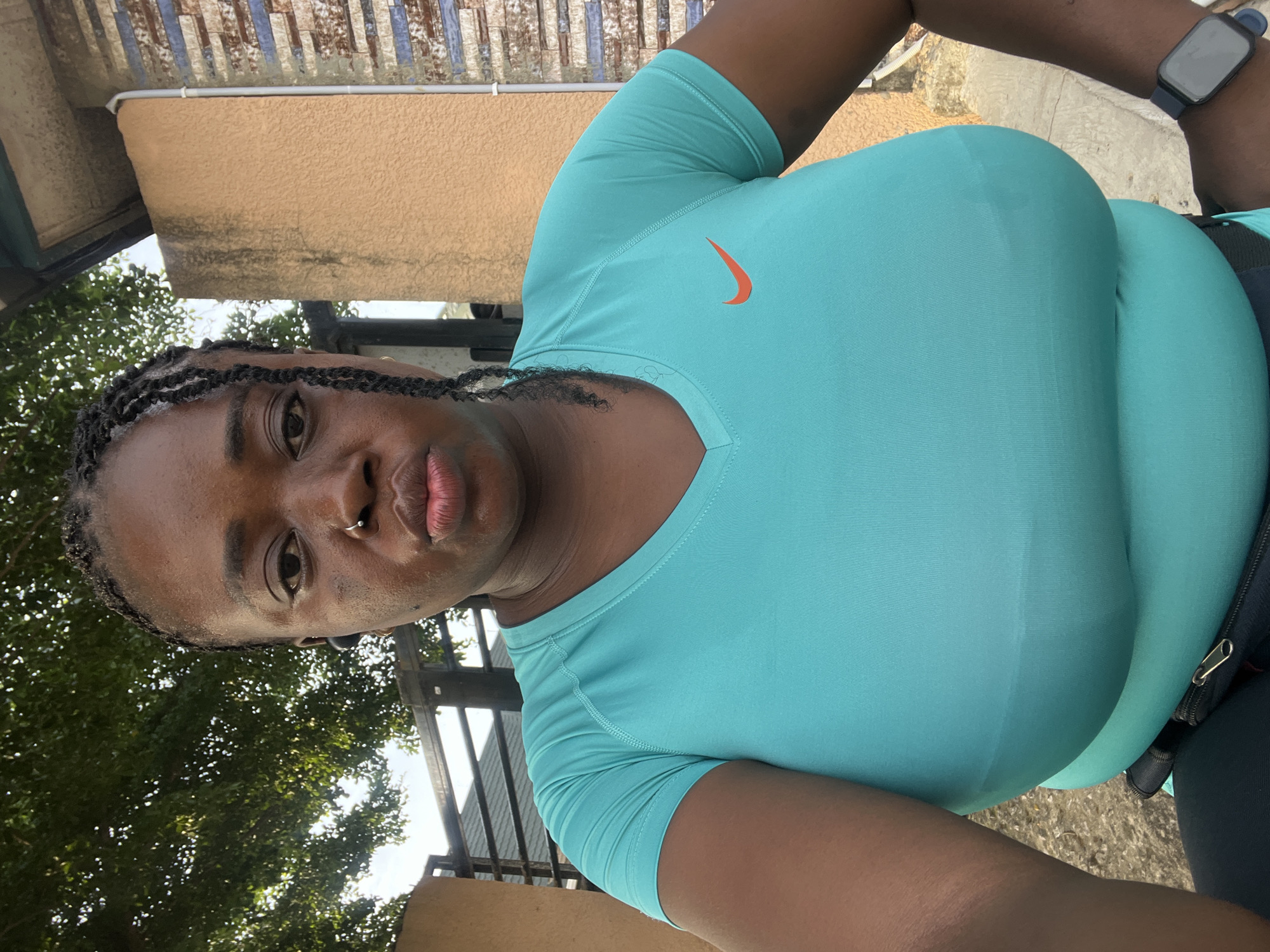 A medium shot captures a woman with dark skin and braided hair, wearing a teal Nike t-shirt and black leggings. She has a nose ring and earbud in her left ear. She is standing outdoors in what appears to be an urban environment, with a beige wall and a section of patterned tiling visible in the background. A large tree with green foliage is to her left, and a metal fence is also present. The lighting suggests it is daytime.