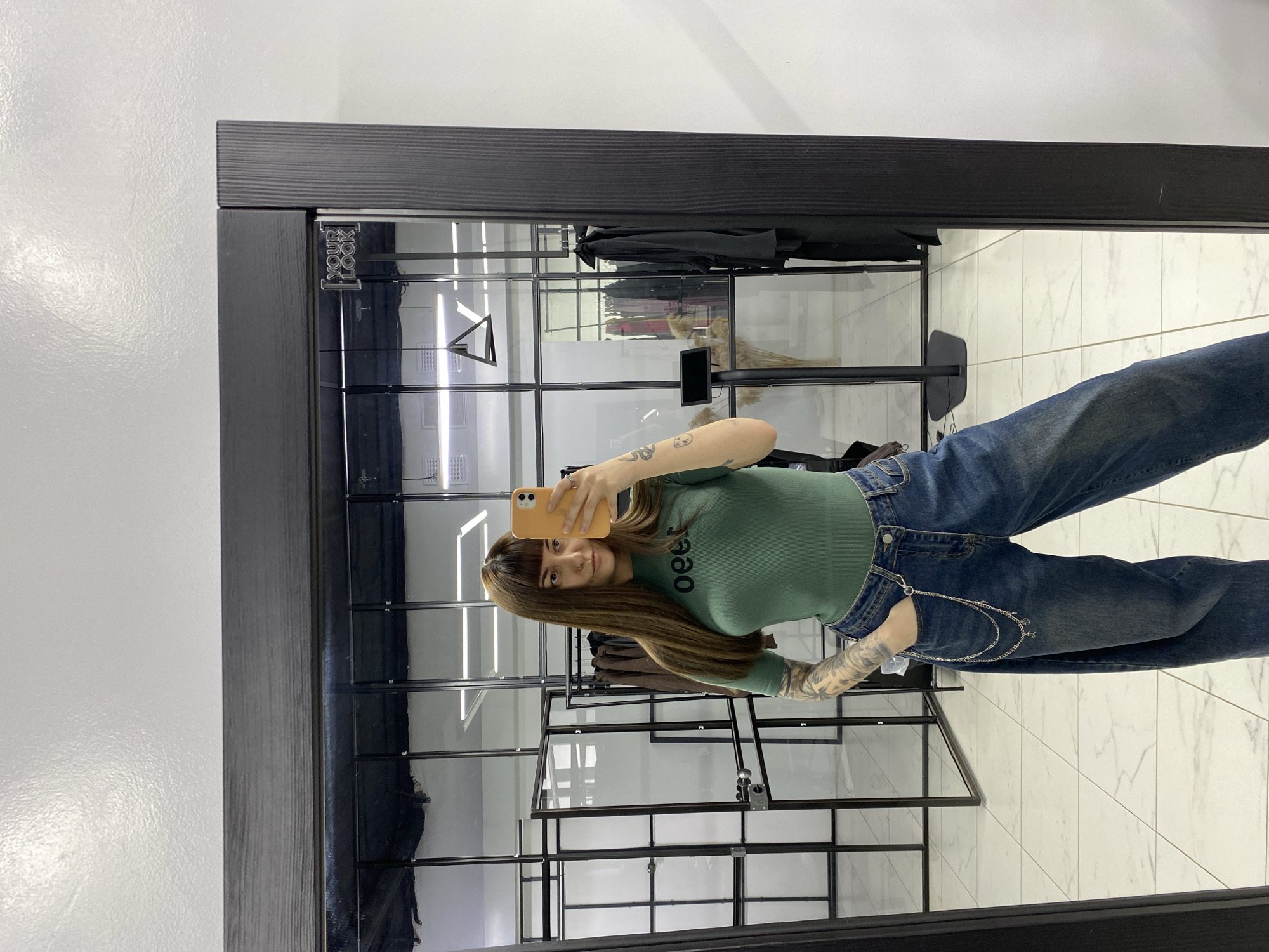 A young woman is taking a selfie in what appears to be a clothing store. She is wearing a green t-shirt, blue jeans with a silver chain, and has long brown hair with bangs. The woman is holding an orange smartphone and looking at the camera. Her left arm, visible in the mirror, has tattoos. The background consists of a black metal-framed clothing rack filled with items, likely clothes, and a white tile floor. The image is taken from the perspective of the mirror's reflection. The location seems to be an indoor retail space. It is impossible to confirm if the store is located in Votkinsk, Russia, from the provided image.