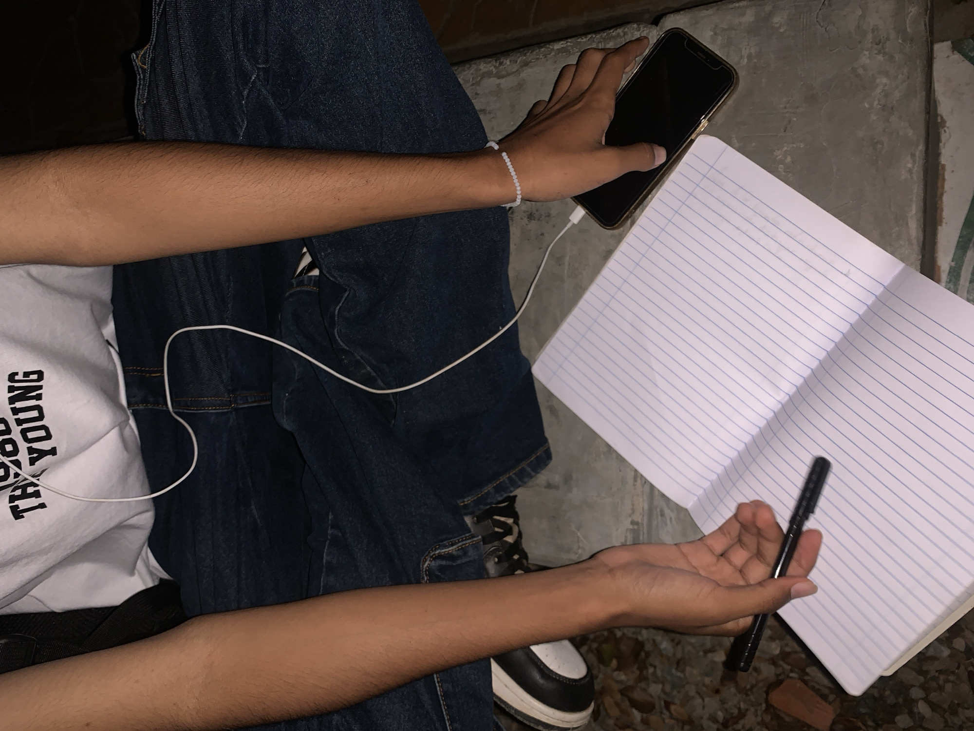 A person sits with a notebook and pen in their hands, suggesting they are about to write. The person wears dark denim jeans and a white T-shirt with black text. A smartphone is connected to the notebook via a charging cable. The background shows the ground with pebbles and a concrete surface. It is unknown if the location is Juan Griego, Venezuela.