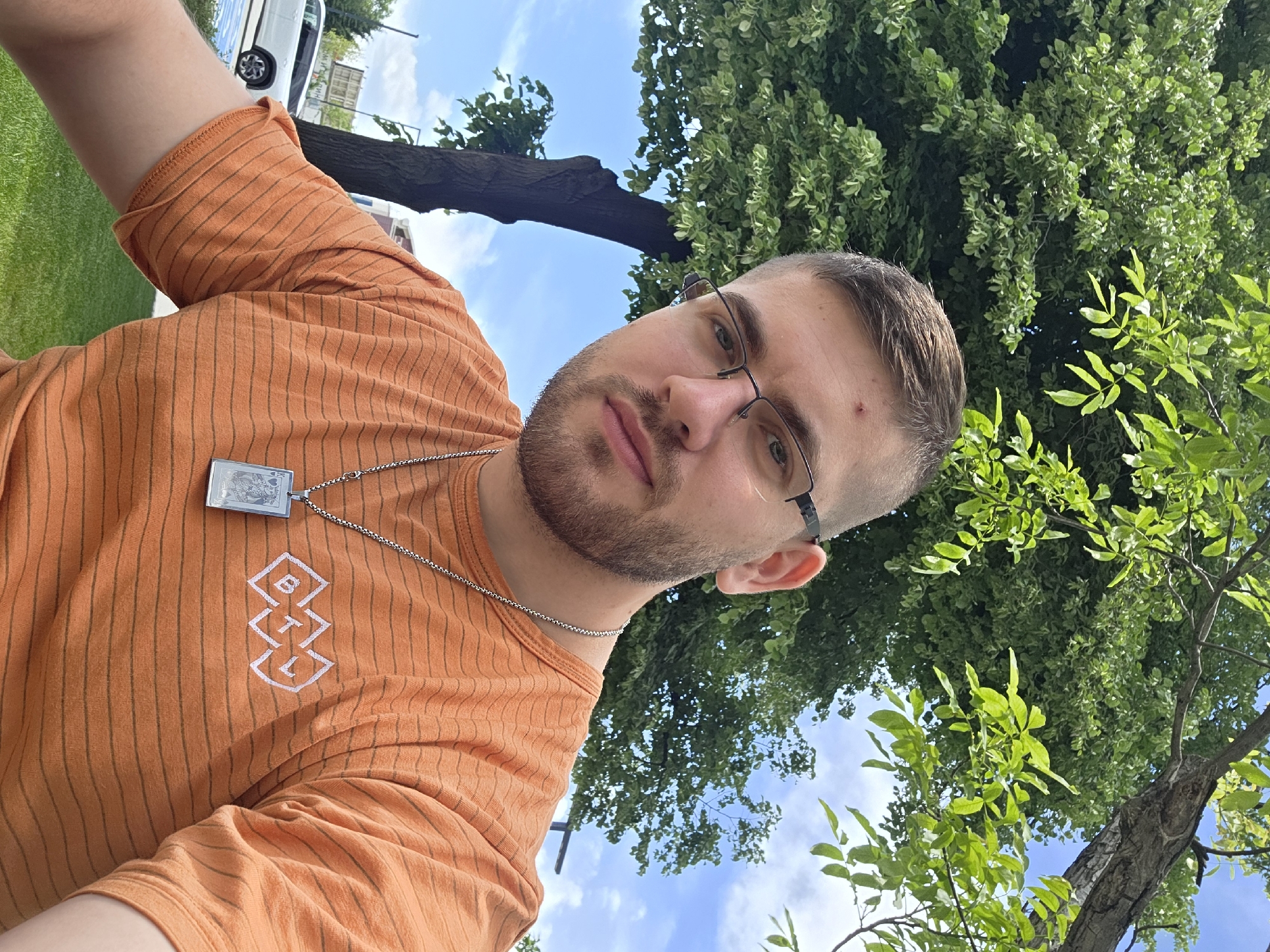 A man is pictured from a low angle, looking at the camera. He has a beard and is wearing glasses, an orange striped t-shirt with a white logo, and a silver necklace with a rectangular pendant. Behind him, there's a view of green trees, a blue sky with white clouds, and part of a white vehicle. The location is likely a park or green area in the city of Plovdiv, Bulgaria, given the presence of greenery and urban elements.