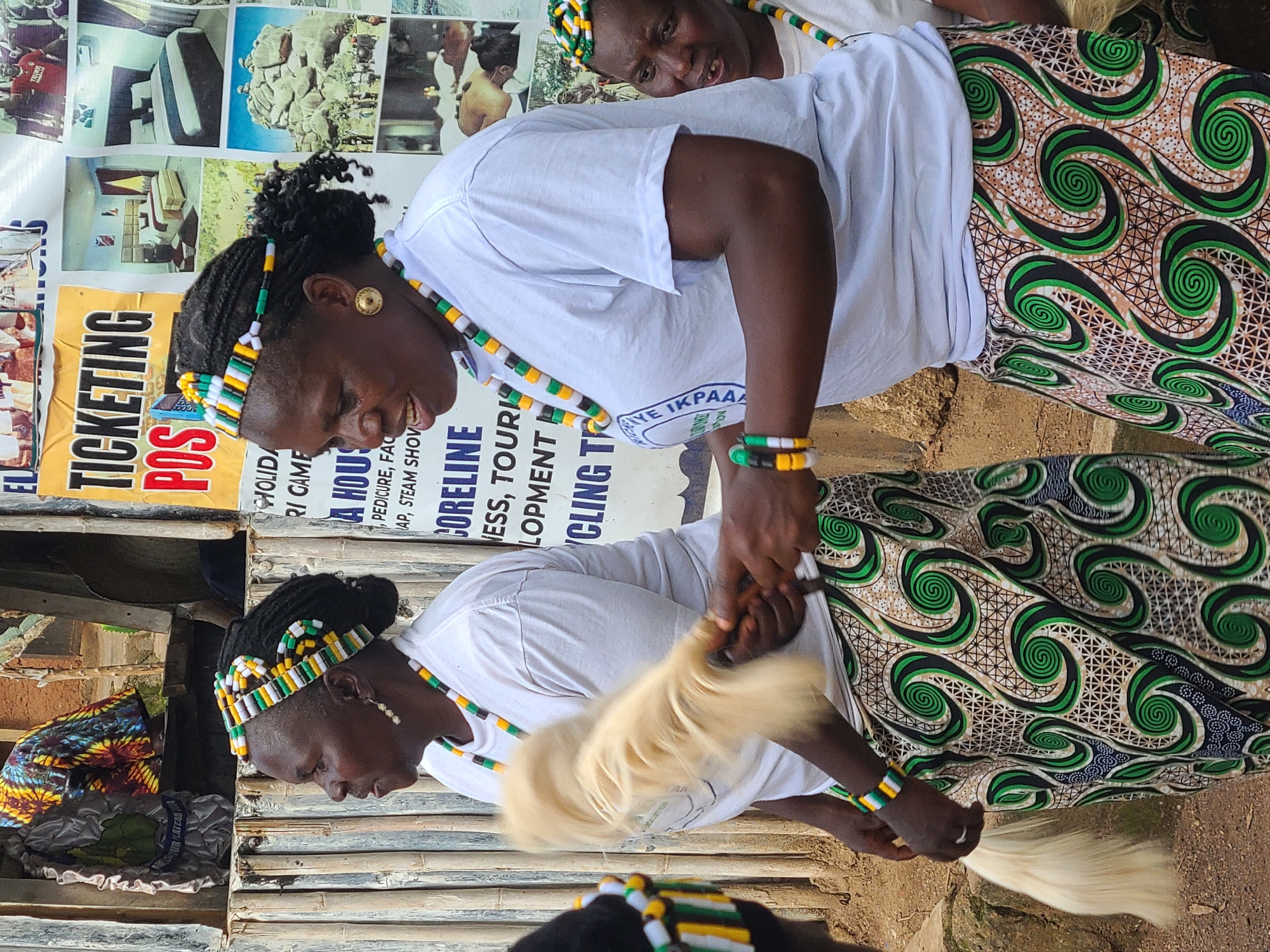 In Jos, Nigeria, two women in traditional attire stand in front of a bamboo stall adorned with colorful fabrics and posters. The woman on the left, seen from the side, wears a white t-shirt, a vibrant patterned skirt, and a beaded headband. She holds a light-colored, fluffy object, possibly a fly whisk, in her right hand. The woman on the right, facing the camera, also wears a white t-shirt, a similar patterned skirt, and a matching beaded headband and necklace. She has her right hand raised, holding the fly whisk, which is in motion, appearing blurred. Both women have elaborate beaded headbands and necklaces. Behind them, posters with text and images are displayed, including pictures of hotel rooms and a large rock formation. The environment appears to be an outdoor market or roadside stall, with natural light illuminating the scene. Another woman is partially visible on the far right, also wearing similar beaded accessories.