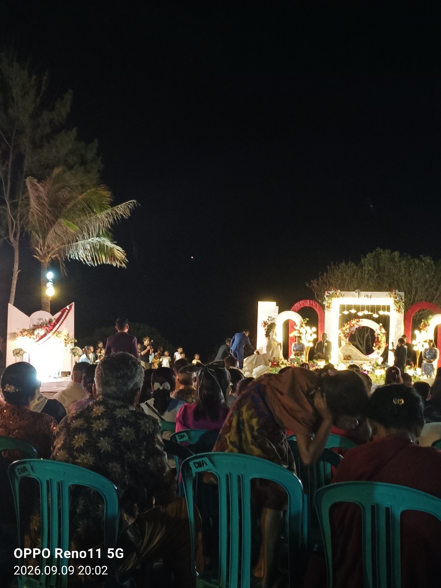 This image depicts an outdoor evening event, likely a wedding reception, in Otan, Indonesia. In the background, a large illuminated structure spelling out "HANI" is visible, adorned with floral decorations and lights, serving as a backdrop for a stage area. Several individuals, possibly a wedding party, are present on and around the stage. The foreground is filled with an audience seated in rows of teal chairs, facing the stage. The scene is illuminated by the stage lights and ambient light, with the dark night sky above. Palm trees and other foliage are visible to the left of the frame. A timestamp overlay in the lower left corner reads "OPPO Reno11 5G 2026.09.09 20:02".