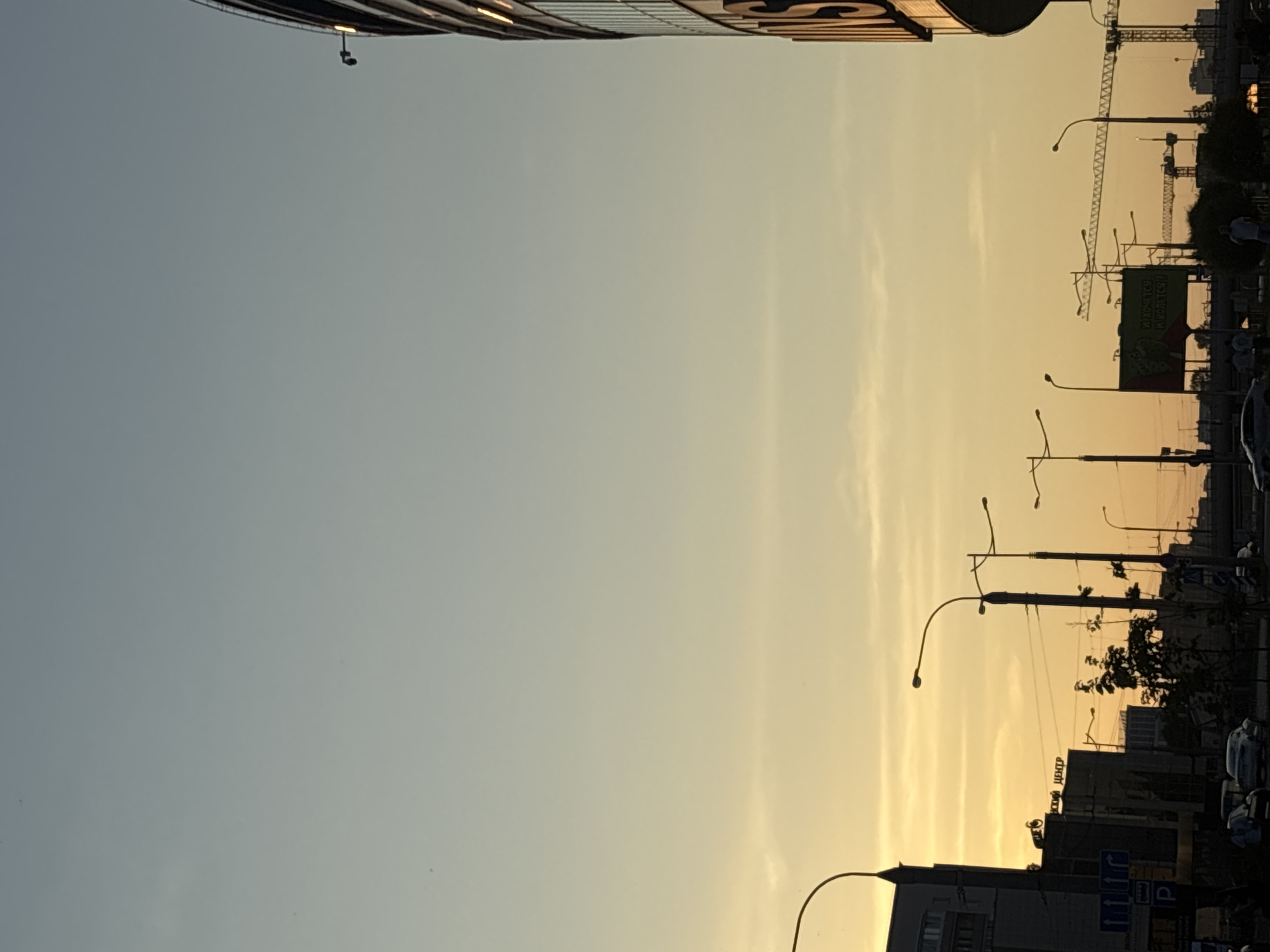 The image captures a city street scene in Minsk, Belarus, during a sunset. The sky is a gradient of pale blue at the top transitioning to warm yellow and orange hues near the horizon. Silhouetted against the sky are several streetlights with curved arms, some appearing more prominent than others.
On the left side of the frame, a tall, modern building with dark windows is partially visible, featuring some illuminated sections. Adjacent to it, another building displays text, with the word "ЦЕНТР" (Center) clearly legible. A street sign with directional arrows and a "P" symbol for parking is also visible.
In the mid-ground and background, a series of streetlights and power lines crisscross the scene. Cranes, indicative of ongoing construction, rise above the lower structures. Several vehicles are present on the road, including cars, some of which are driving. A few individuals are discernible as small figures walking or standing.
On the right side of the image, a large, curved building with a reflective facade dominates the edge of the frame. Illuminated lights are visible on its upper floors, and a large, dark, vertical sign with partial lettering is attached to its side, suggesting it is a prominent establishment. The overall impression is an urban environment at dusk, with the warm light of the setting sun casting long shadows and highlighting the architectural features of the city.