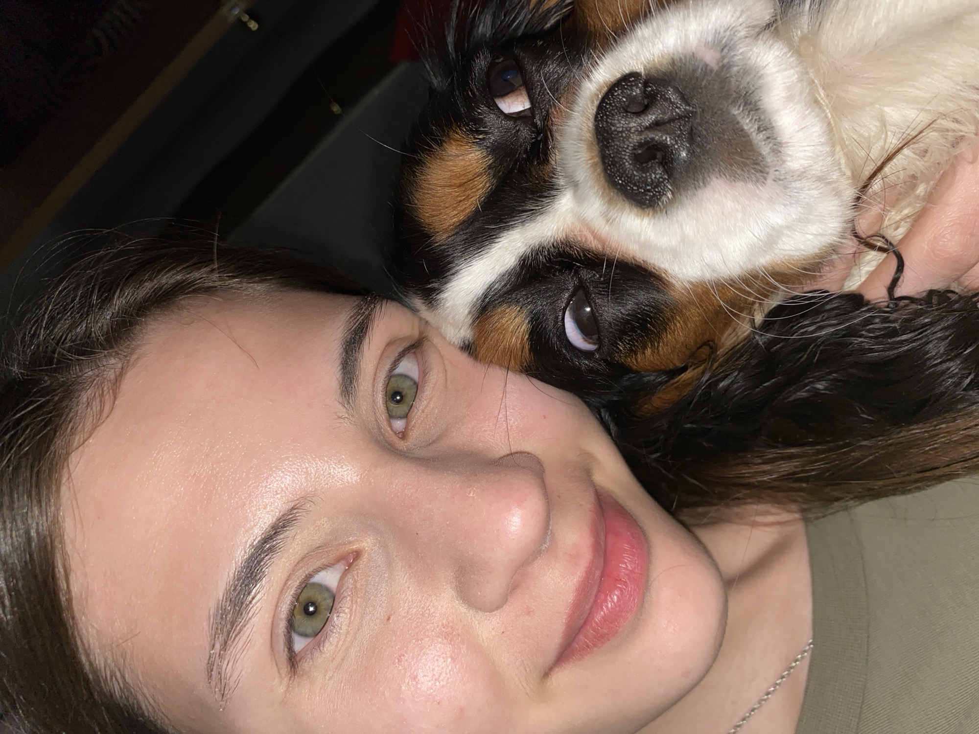 In this close-up selfie, a young woman with light skin, dark brown hair, and green eyes is pictured with a dog. The woman is looking at the camera with a slight smile. The dog, a Cavalier King Charles Spaniel with tri-color markings, is positioned next to the woman's face, with its head tilted and eyes looking upwards, giving it a slightly comical expression. The background is dark and out of focus, making the subjects the clear focus of the image. There are no discernible details of Votkinsk, Russia in the image.