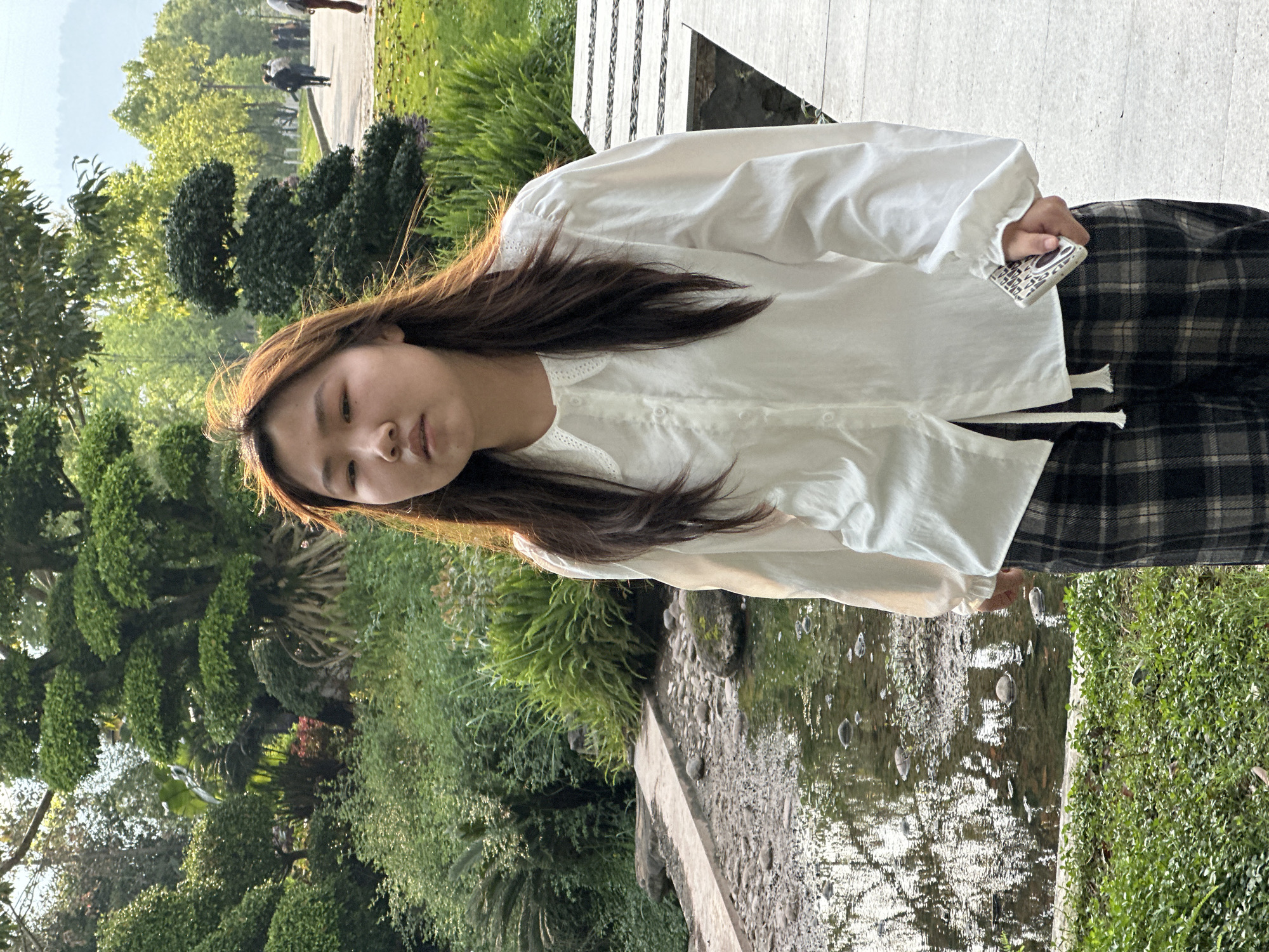 A woman stands in a park-like setting in Jiangjin, China. She wears a white long-sleeved blouse and a plaid skirt or pants. In her hand, she holds a small, rectangular object with a patterned surface. The background features lush greenery, trees, a small body of water, and a walkway. Sunlight illuminates the scene, casting shadows and highlights.