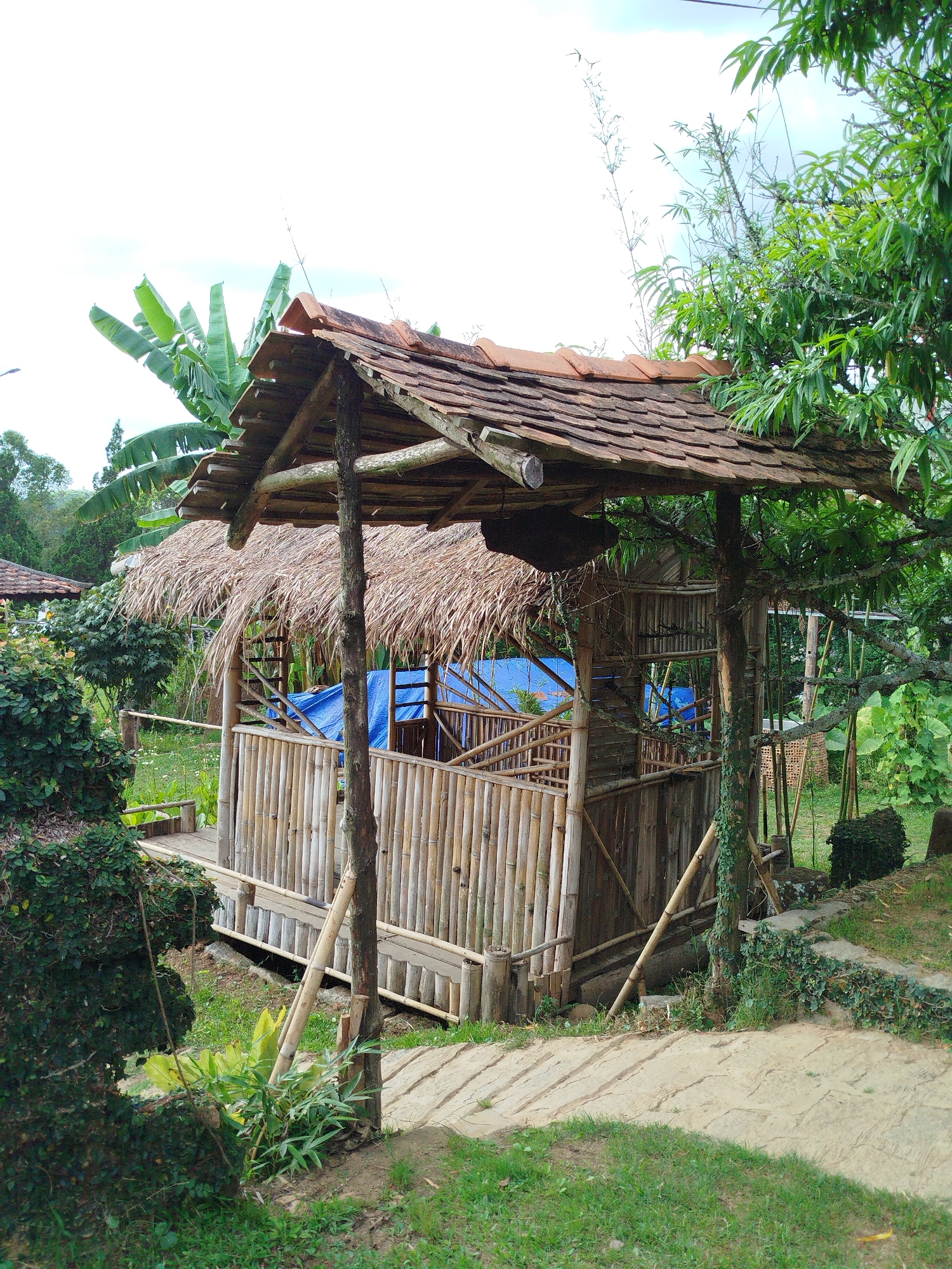 An outdoor view shows a traditional wooden and bamboo structure resembling a small pavilion or gazebo. The structure has a thatched roof with a layer of clay tiles on top. The sides are constructed of bamboo, with some sections covered by a blue tarp. It is supported by wooden posts and situated in a garden setting with green grass and various plants and trees. A path leads to the structure. The location is suggestive of a natural or tourist setting, potentially within the city of Da Lat, Vietnam.
