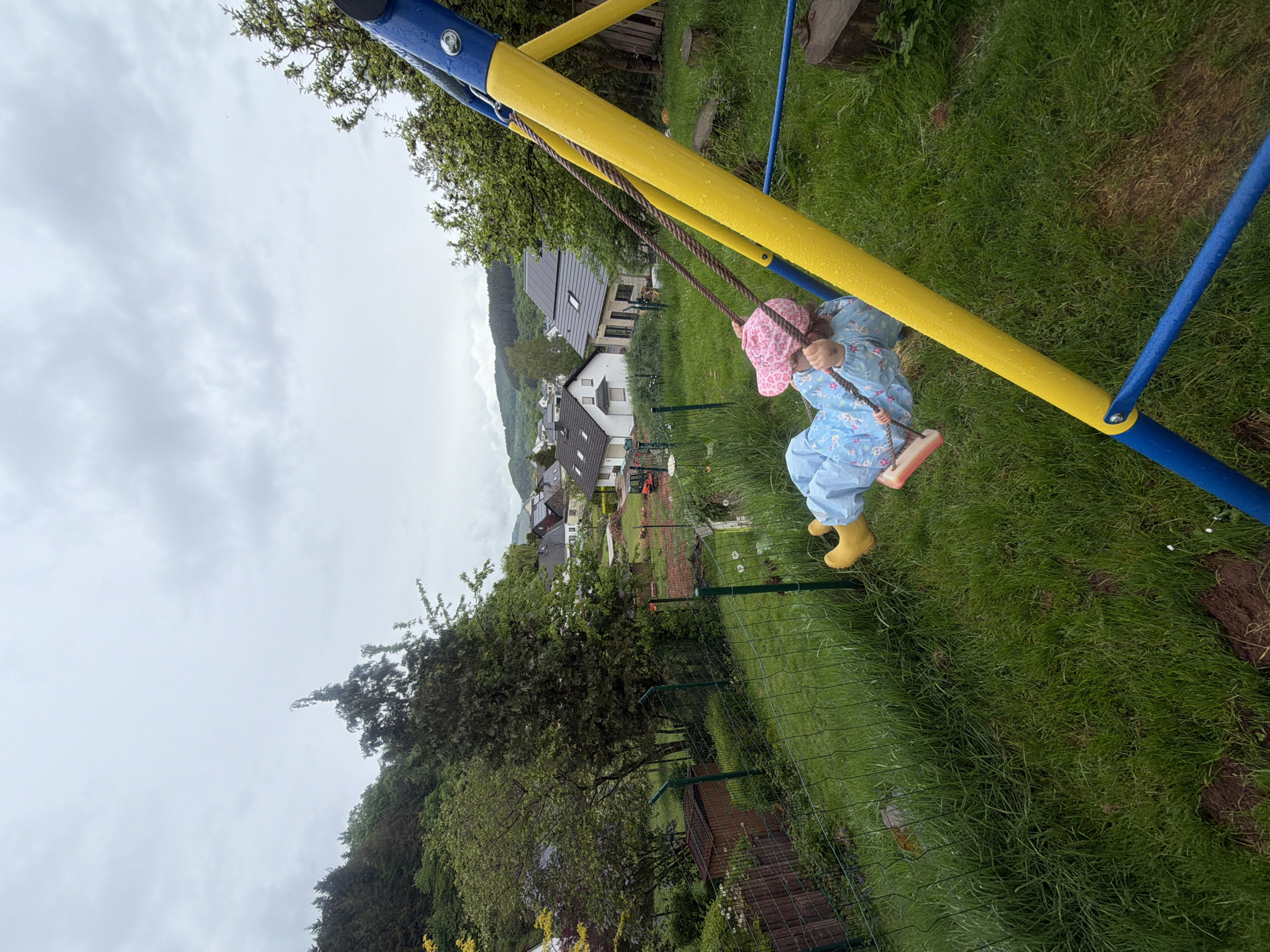 A young child wearing a pink patterned hat, a blue patterned raincoat, and yellow boots is on a swing. The swing is part of a yellow and blue metal swing set. The environment is a grassy yard with a wire fence. In the background is the town of Trassem, Germany, featuring houses with varying roof types and colors, surrounded by trees and a slightly overcast sky.