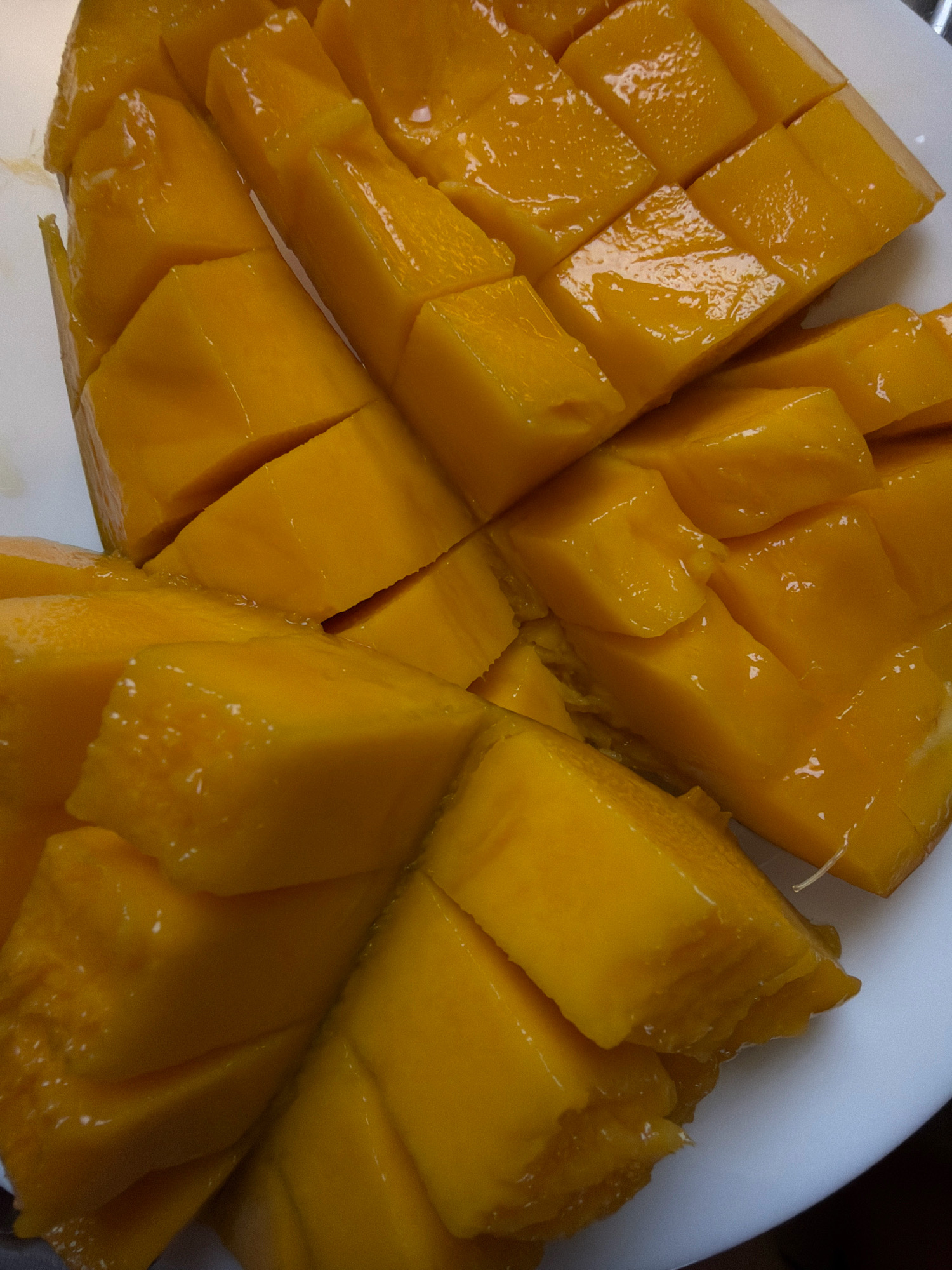 A close-up, top-down shot shows a white plate filled with cut mango pieces. The mango flesh is a vibrant orange-yellow, and the cuts reveal a smooth, juicy texture. Some pieces are arranged in rows, while others are scattered more randomly. The lighting casts subtle shadows, highlighting the curves and angles of the fruit. The overall impression is one of freshness and abundance. The image is likely taken indoors, in a kitchen or dining area. There is no discernible urban landscape or any indication of the city of Sharm El Sheikh, Egypt, or any specific location within it.
