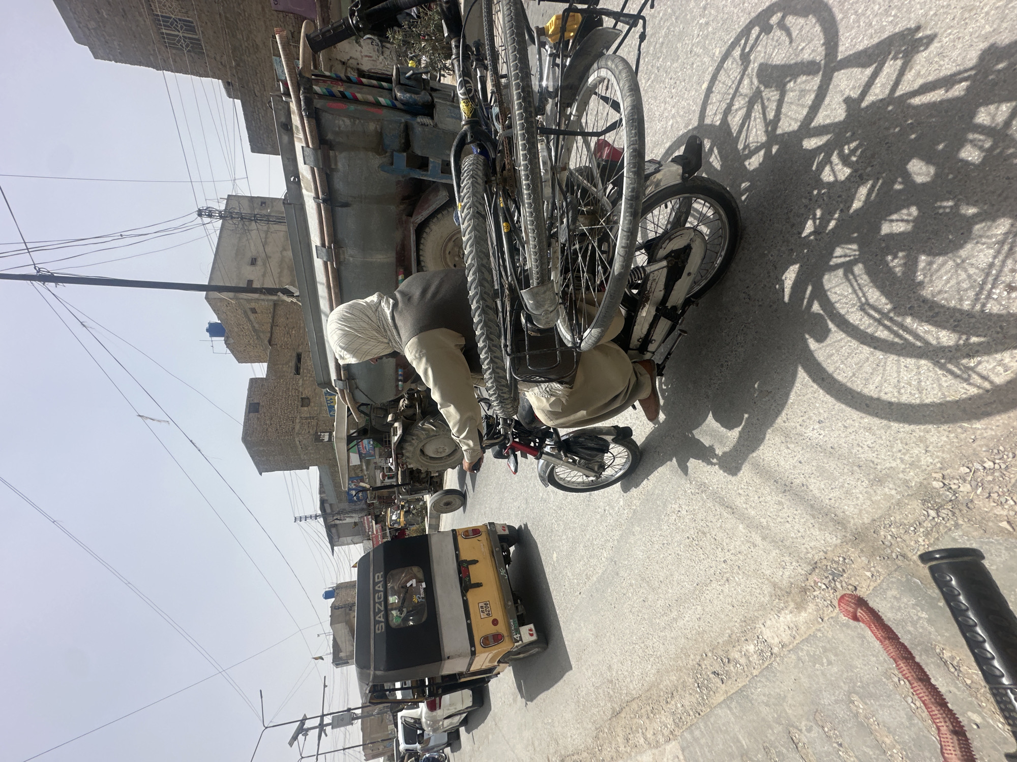The scene appears to be an urban street in Quetta City Tehsil, Pakistan. An individual, wearing a light-colored head covering, is kneeling, apparently working on a small motorcycle attached to a larger truck or tractor trailer carrying bicycles. The vehicles are positioned on an asphalt road. In the background, there is a yellow and black auto rickshaw, and other unidentified vehicles. Utility poles and power lines are visible overhead, as are brick and concrete buildings. The scene is bathed in bright sunlight, creating sharp shadows on the ground.