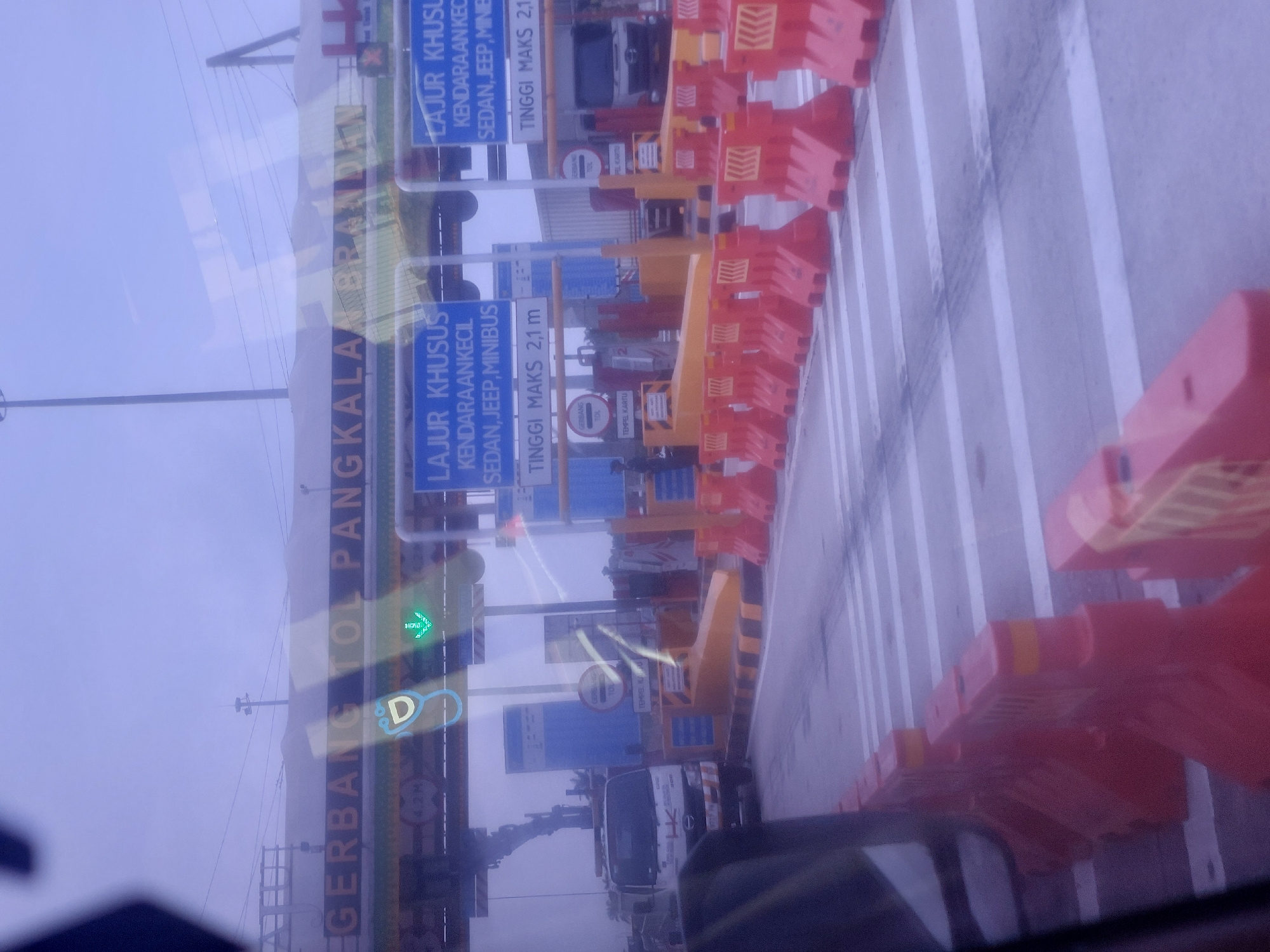 The image depicts a toll plaza on an overcast day, likely in Tangkahan Durian, Indonesia, as indicated by the signage. The foreground is dominated by a line of bright orange traffic barriers, delineating multiple toll lanes. Beyond the barriers, several toll booths are visible. Each booth features overhead signage in Indonesian. One sign reads "GERBANG TOL PANGKALA BRANDA," identifying the toll gate. Below this, blue signs specify "LAJUR KHUSUS KENDARAAN KECIL SEDAN, JEEP, MINIBUS" and "TINGGI MAKS 2,1 m," indicating lanes designated for smaller vehicles and a height restriction. A green arrow illuminated above a toll booth suggests a lane is open. In one lane, a white truck with "HK" branding is stopped, presumably at the toll booth. The overall environment appears to be a functional toll collection point with clear directional and informational signage for motorists.