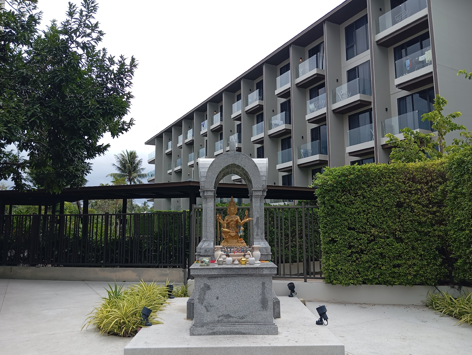The image shows an outdoor shrine in front of a modern building, likely a hotel or condominium, in Karon, Thailand. The shrine is made of gray stone and features a golden statue of a multi-armed deity, possibly from Hindu mythology, set within an arched enclosure. The deity holds various objects in its hands. The shrine is adorned with several small white vases, some small figurines, and what appear to be offerings.
In the foreground, there is a paved courtyard area with a low-lying plant with yellow and green foliage. Black outdoor spotlights are visible around the base of the shrine and along the walkway. Behind the shrine, a black metal fence separates the area from a lush green hedge and a covered walkway. Beyond the fence, trees and palm trees are visible, along with glimpses of the building's structure.
The building itself is several stories high, with a repeating pattern of balconies and windows. The balconies have glass railings and dark frames. The building's facade is painted a neutral gray. The sky is overcast. The scene appears to be a quiet, well-maintained outdoor space, possibly part of a resort or residential complex.