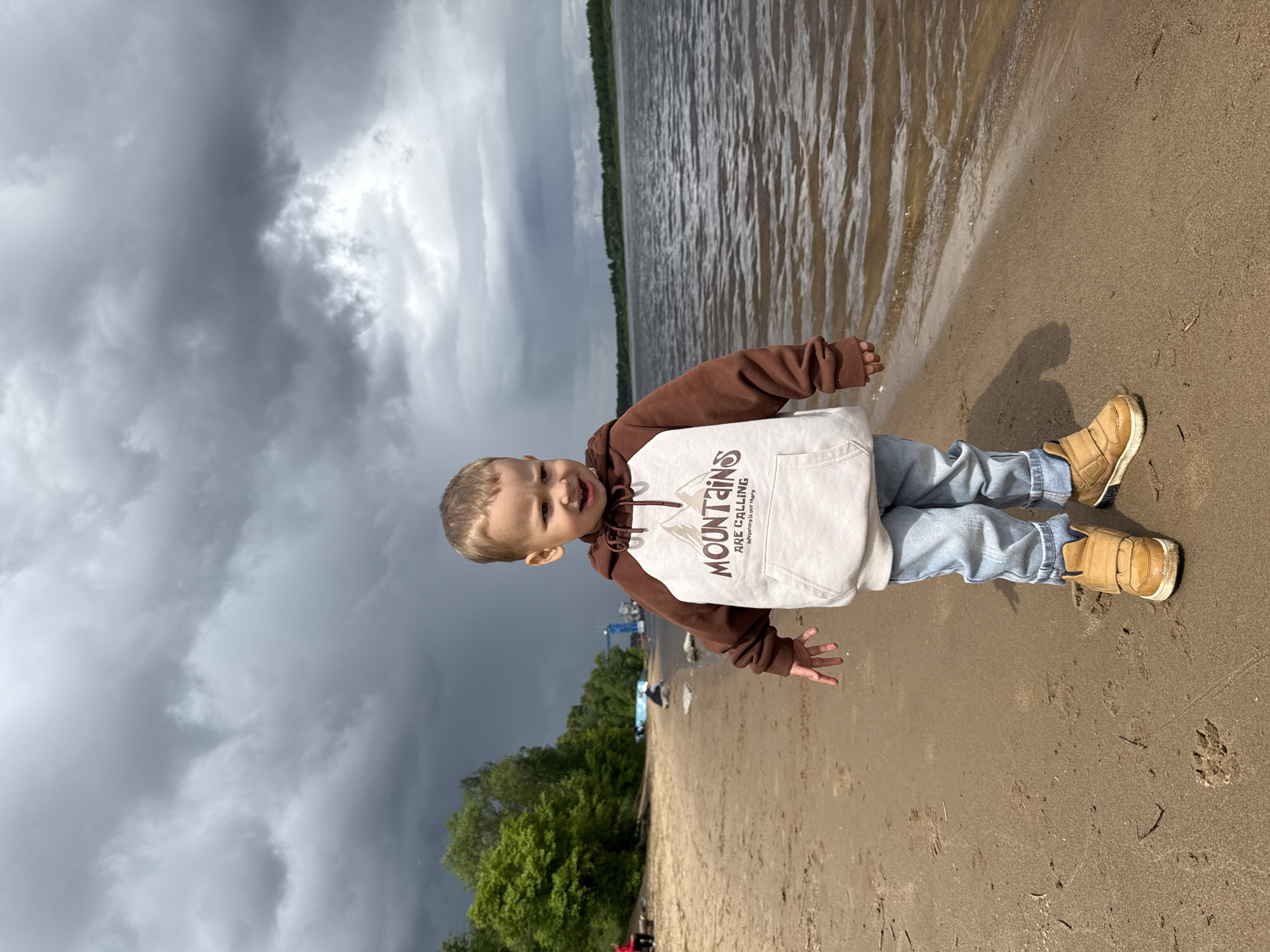 A young child stands on a sandy beach near the edge of a body of water, likely a lake or river, under a dramatic, cloudy sky. The child is wearing light blue jeans, tan shoes, and a hoodie with brown sleeves and a white center with a printed design. The water appears choppy, and trees line the opposite shore. The sky is dominated by dark, ominous clouds, suggesting an approaching storm. The location is near Rusanovo, Russia, according to the prompt, suggesting the presence of the lake, bodies of water, and river.